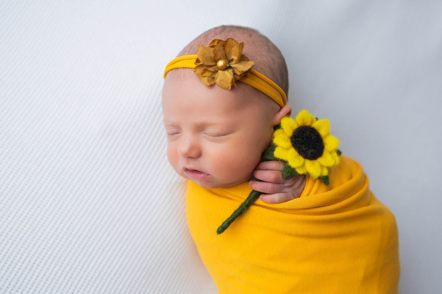 Sleeping newborn wrapped in yellow fabric with sunflowers as decorative accents on white textured background.