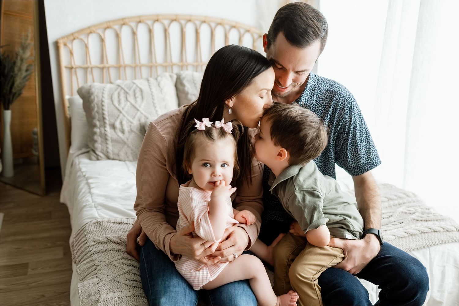 Family of four sitting on bed, parents kissing toddler son and baby daughter in pink bow during studio portrait session.