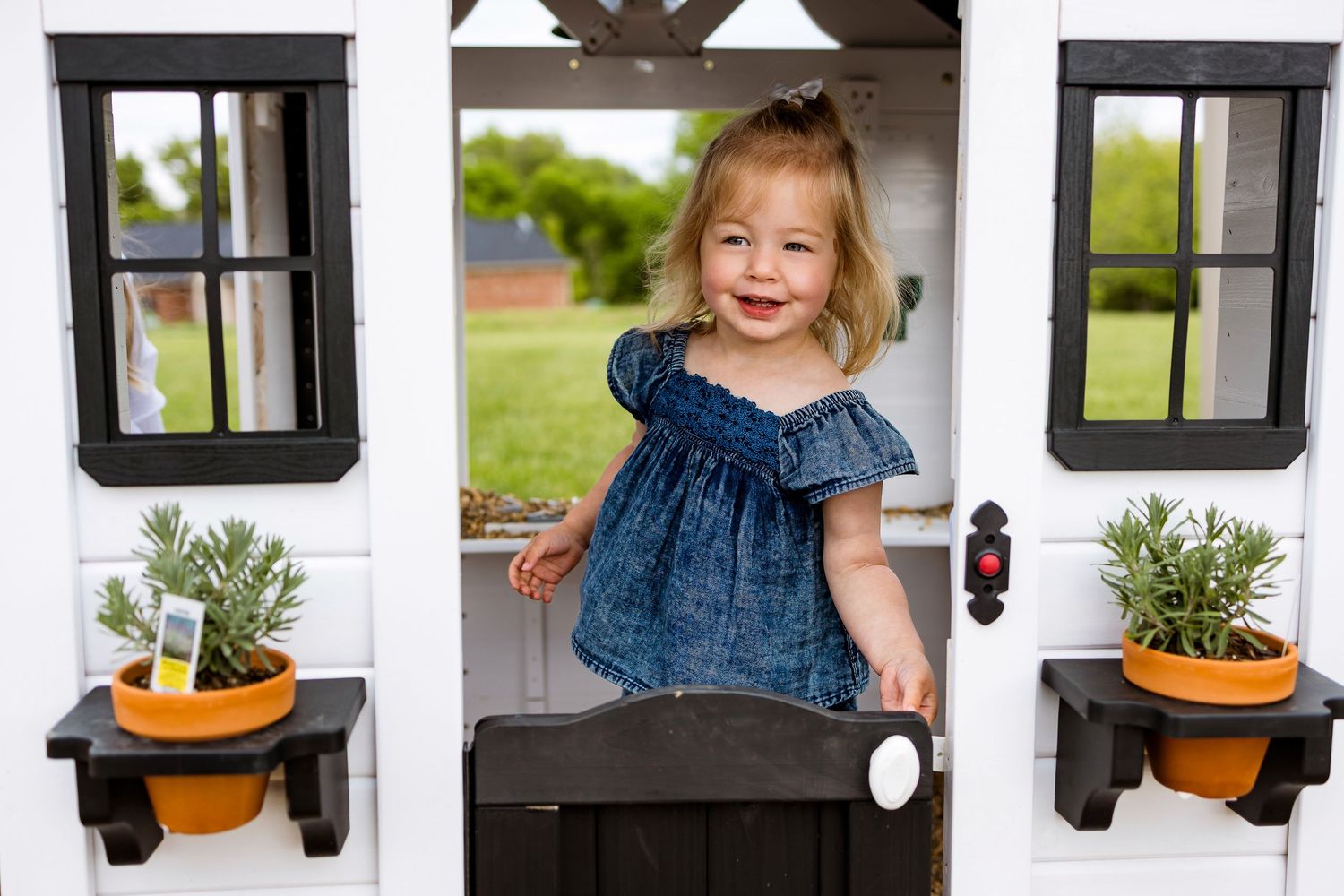 Smiling child in blue dress poses in white playhouse doorway with potted plants.