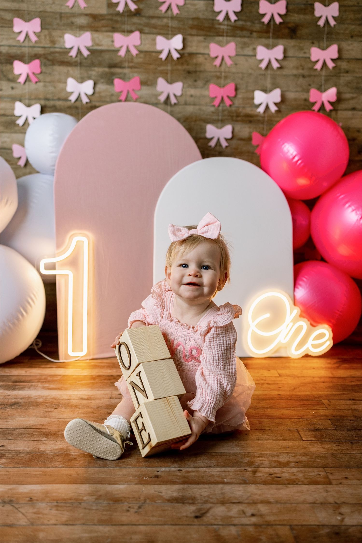 First birthday party scene with pink balloons, decorative bows, and wooden blocks on hardwood floor.