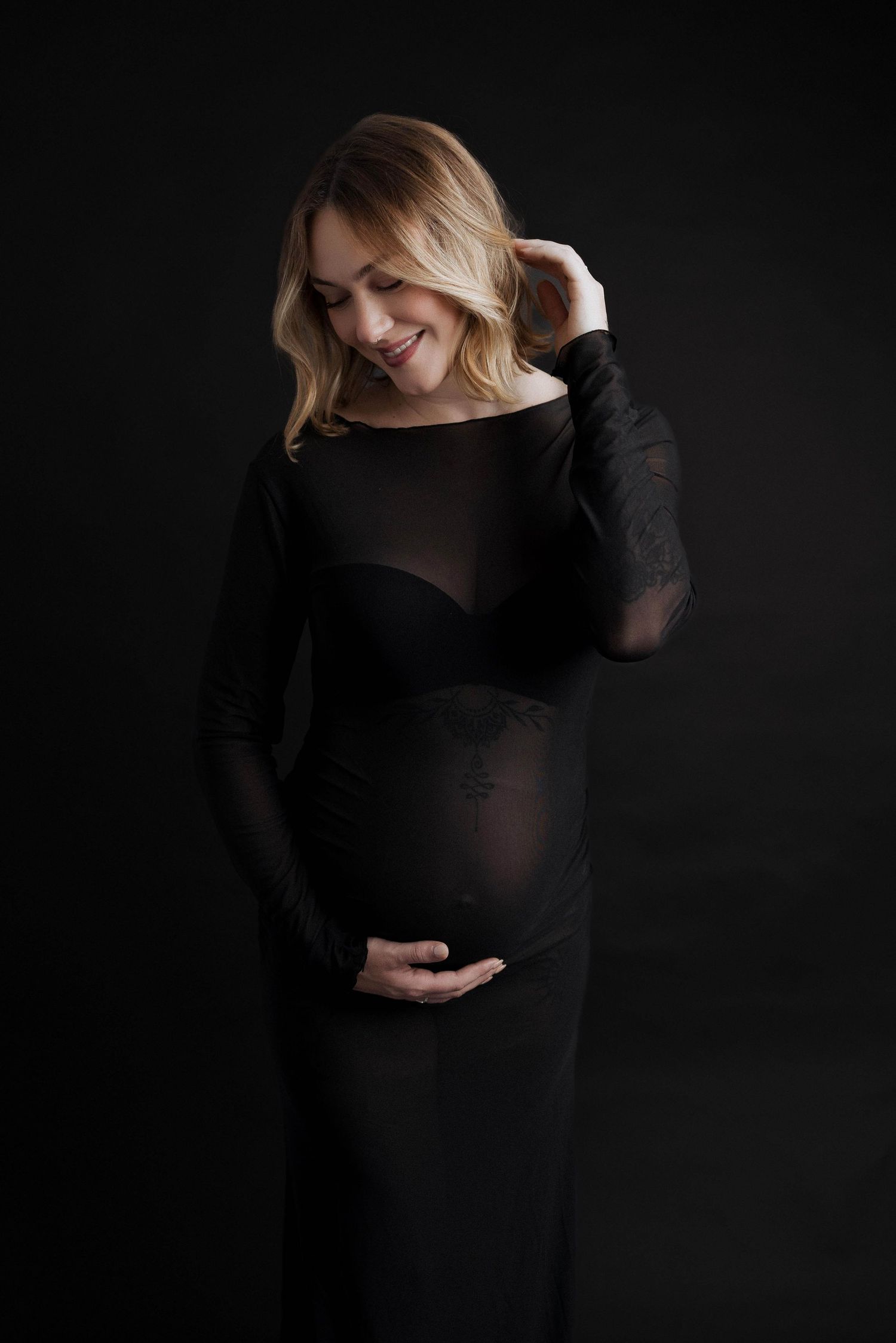 Expecting mother in black dress poses against dark backdrop in dramatic maternity photo.