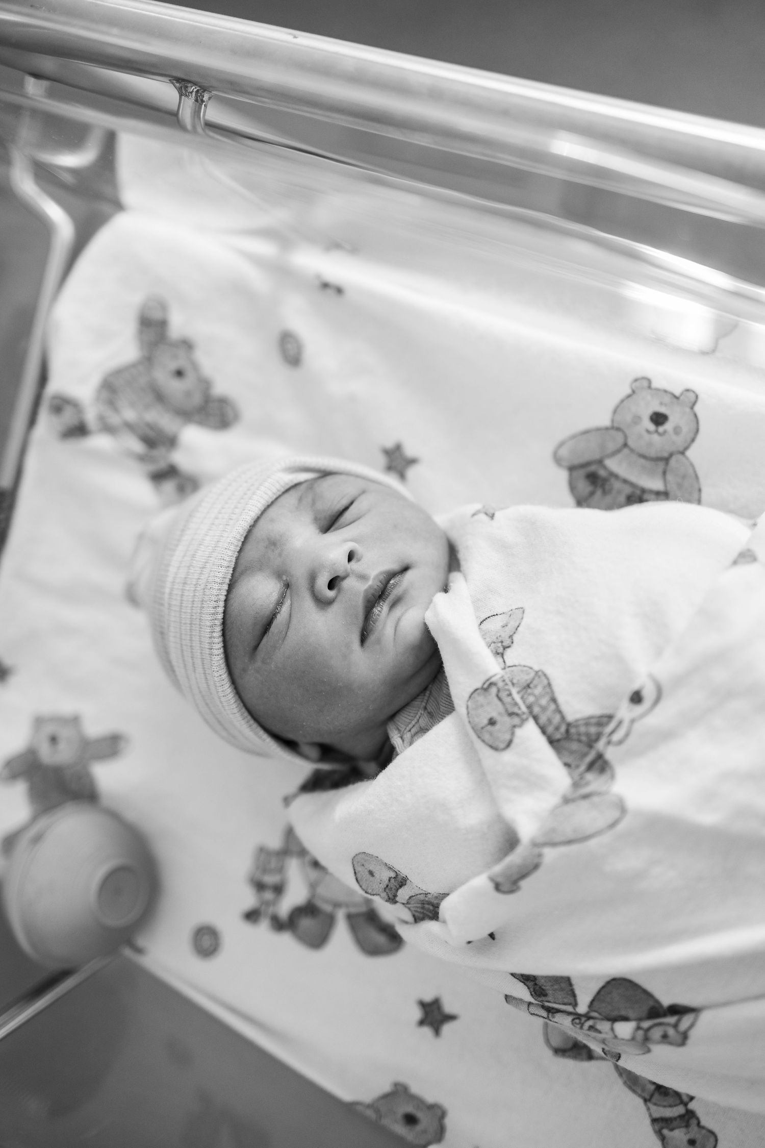 A newborn infant sleeping peacefully in a hospital bassinet with teddy bear printed bedding and wearing a knit cap.