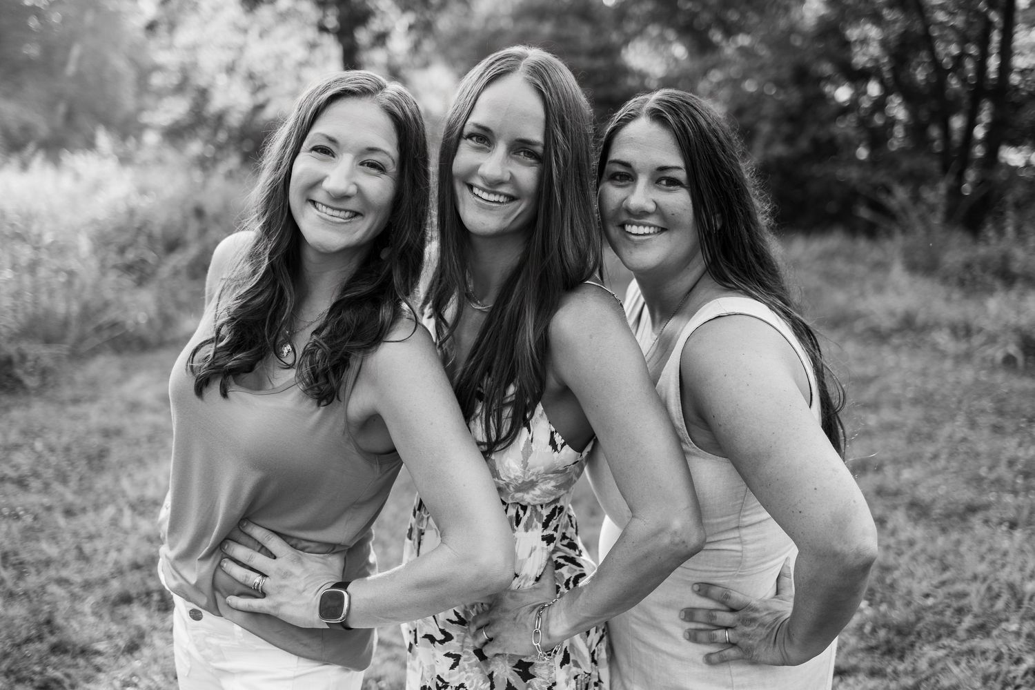 Three women in sleeveless tops smile while posing outdoors in black and white photography.
