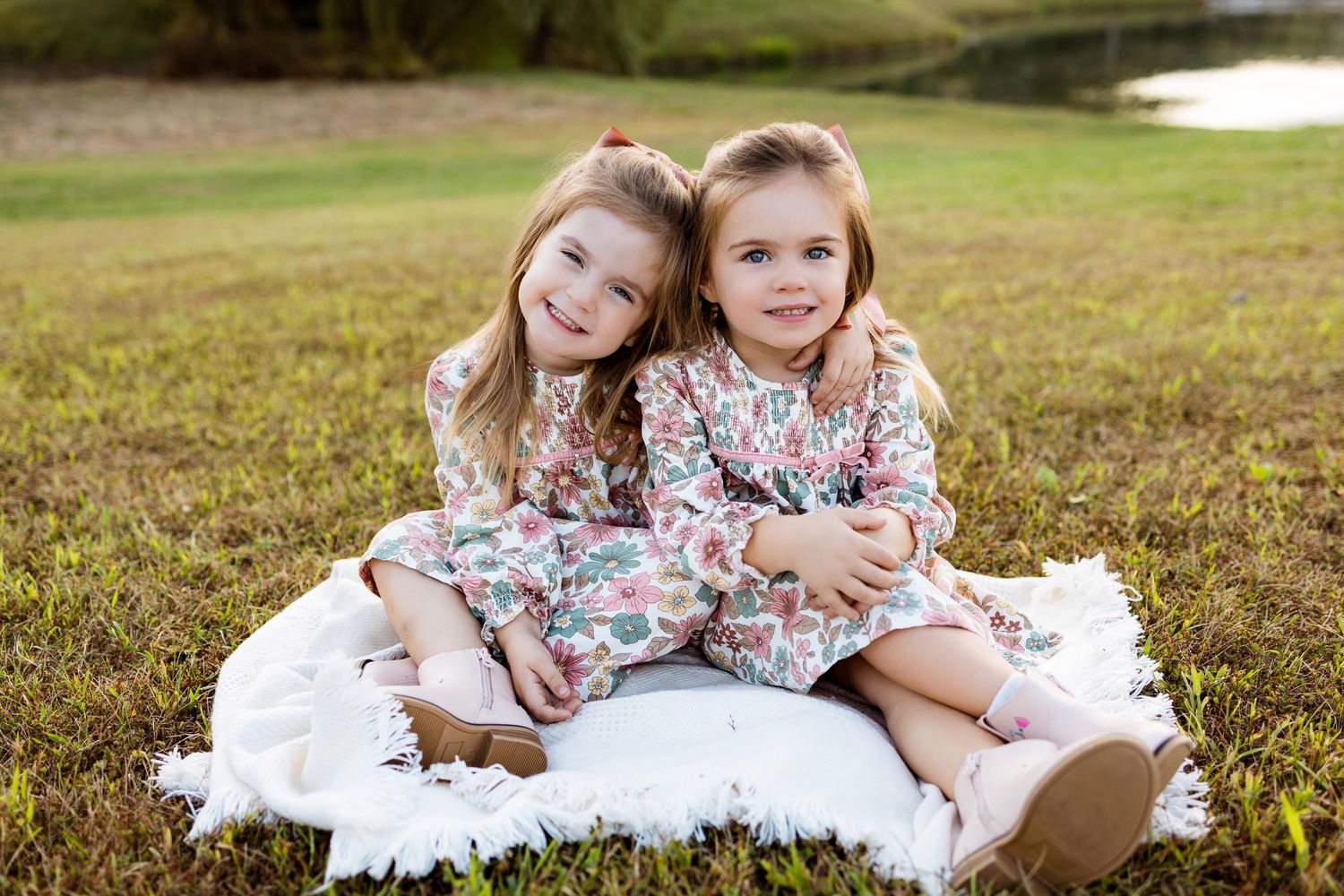 Two children in floral and white dresses sit together on grassy ground during an outdoor photo session.