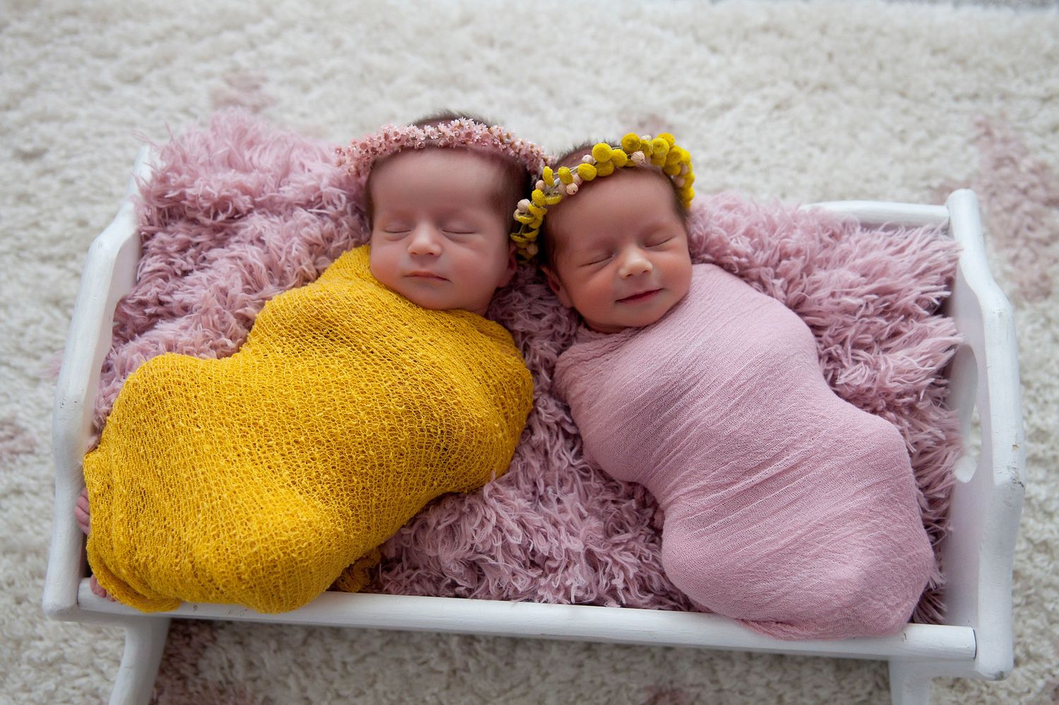Two swaddled newborns wearing knit headbands rest on pink blanket, one wrapped in yellow and one in pink.