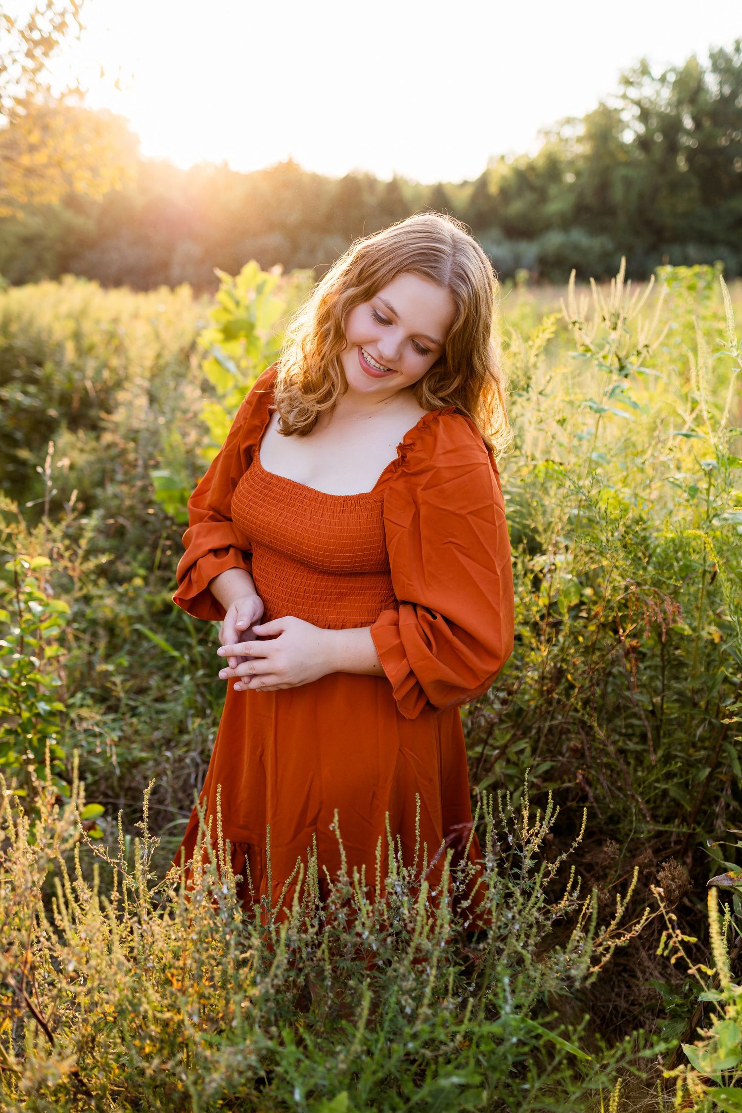 A pregnant woman in a rust-colored dress poses in a golden wildflower field at sunset.
