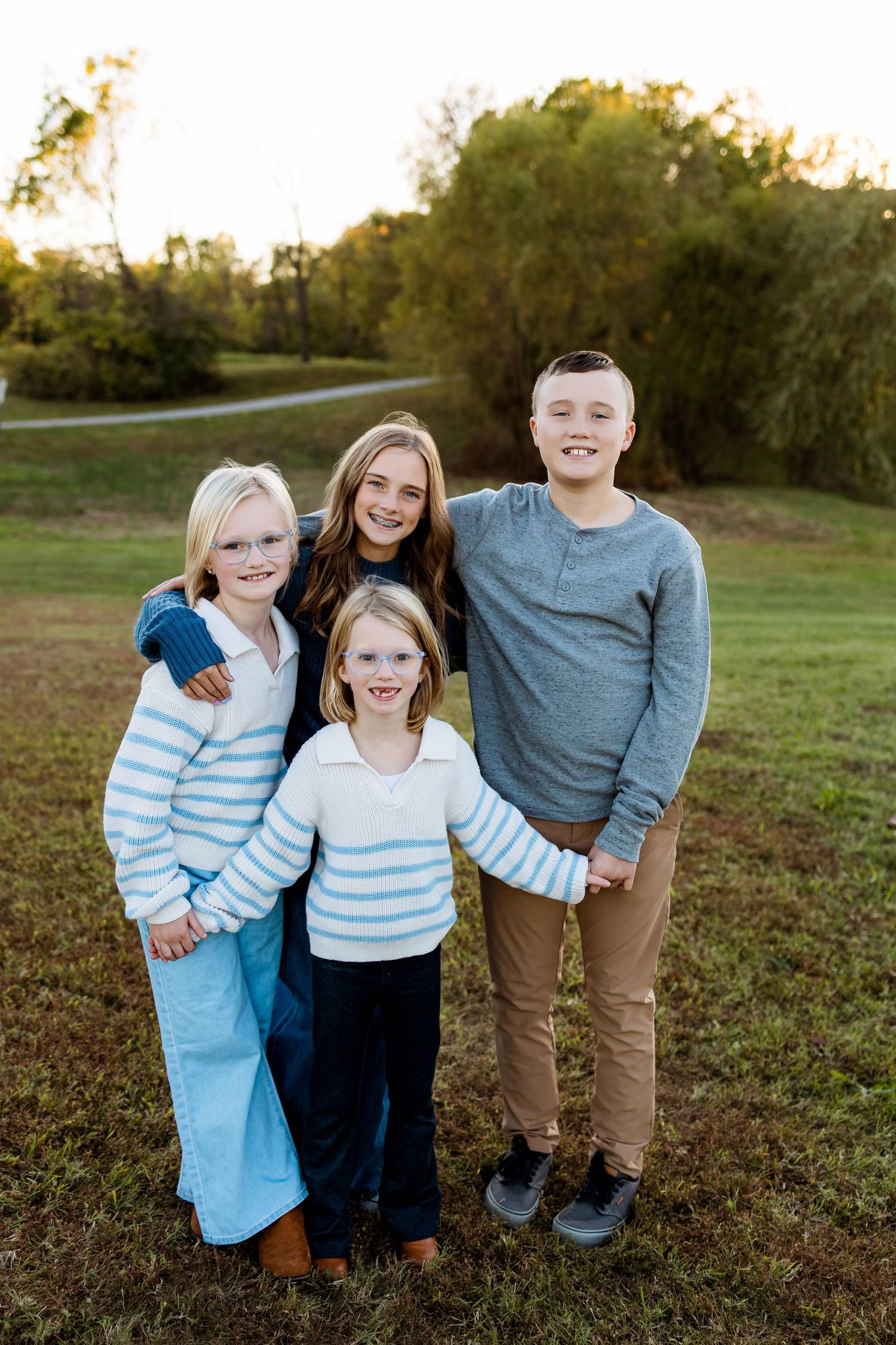 Three siblings pose together outdoors in coordinating blue and neutral outfits on a grassy hillside at sunset.