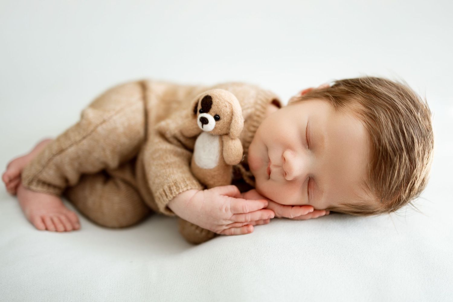 Sleeping baby in brown knit outfit cuddling with plush teddy bear toy on white background.