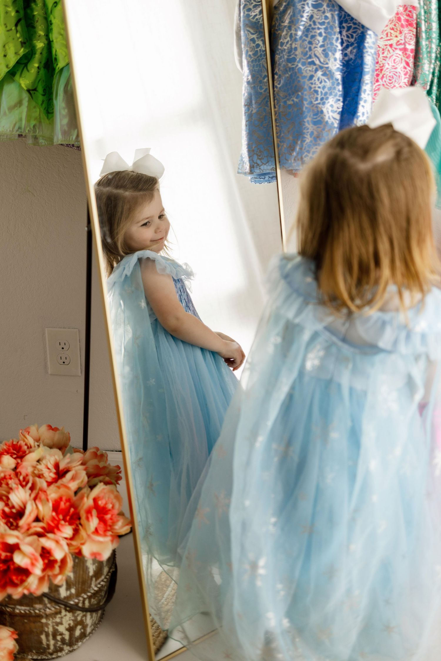 A young child in a light blue princess dress twirls and dances in front of a mirror with floral decorations.