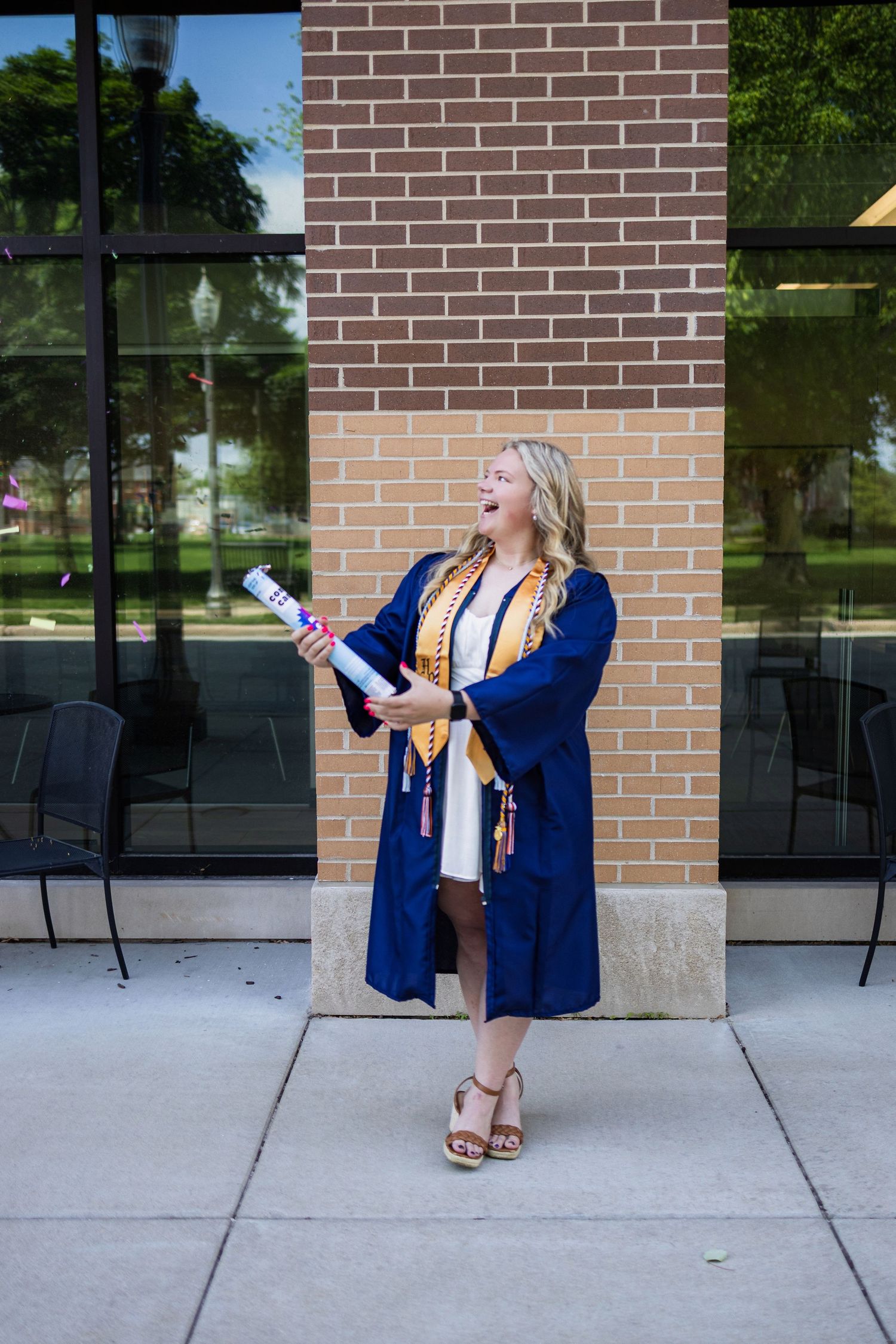 Graduate wearing blue graduation gown poses joyfully with diploma roll in front of brick building.