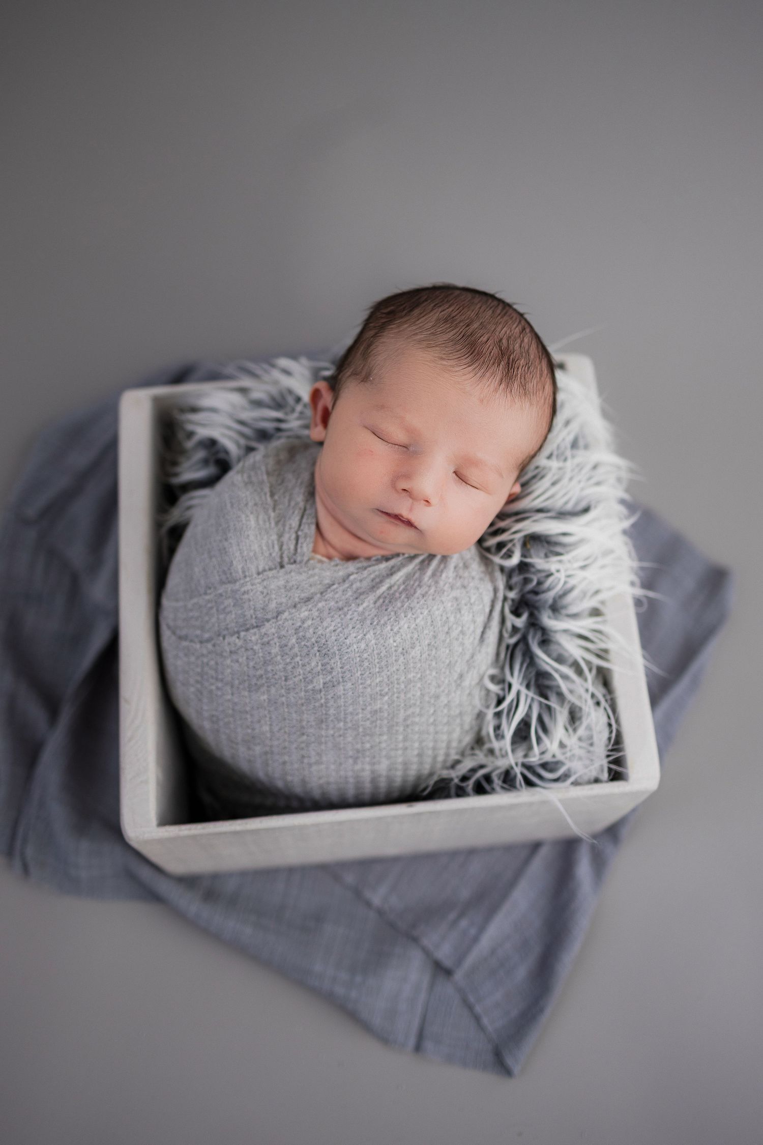 A sleeping newborn wrapped in soft grey swaddle rests peacefully in a white box lined with fuzzy grey fur.