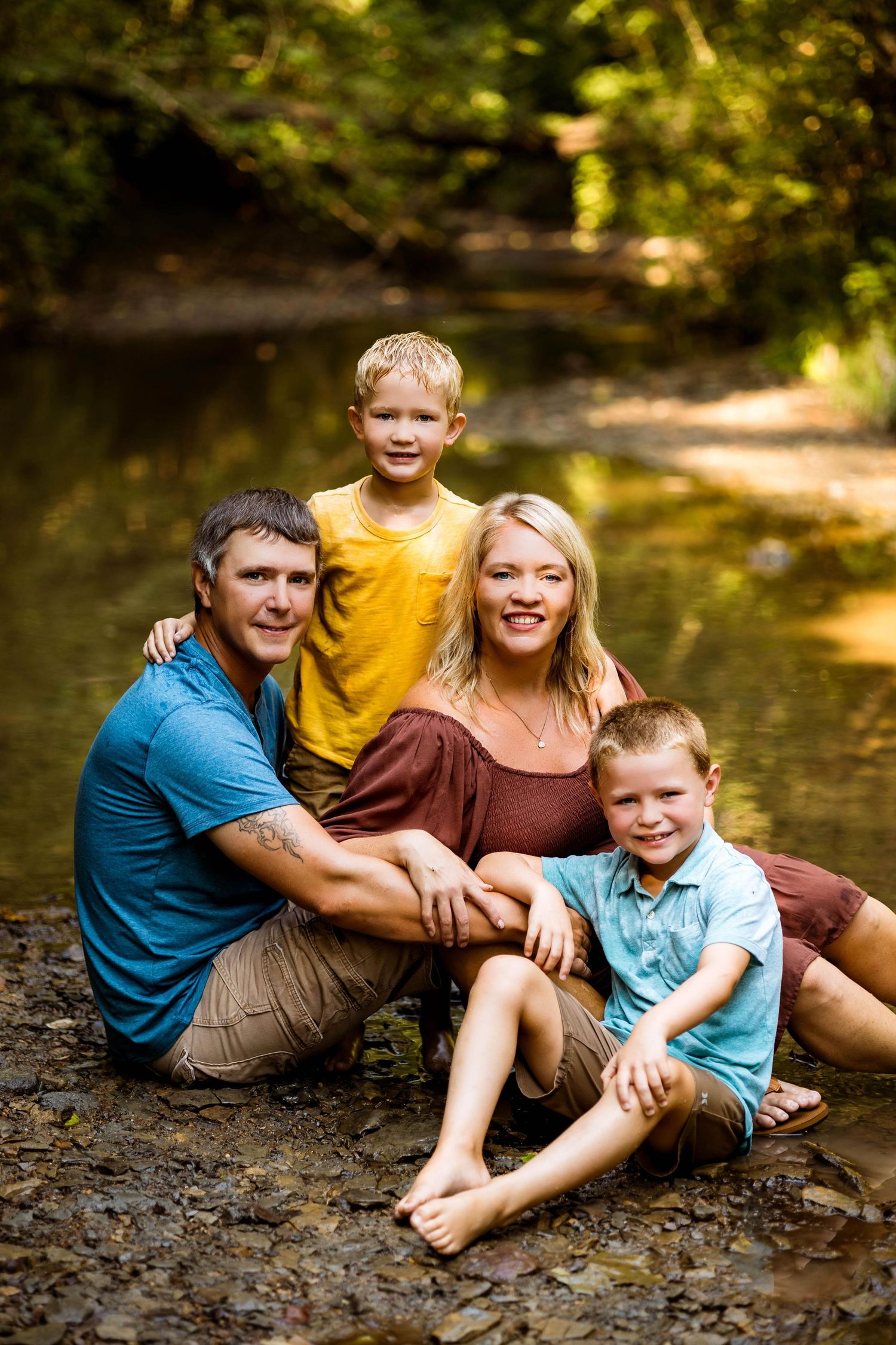 A family shares joyful laughter and embraces while sitting on rocks near a creek in dappled sunlight.