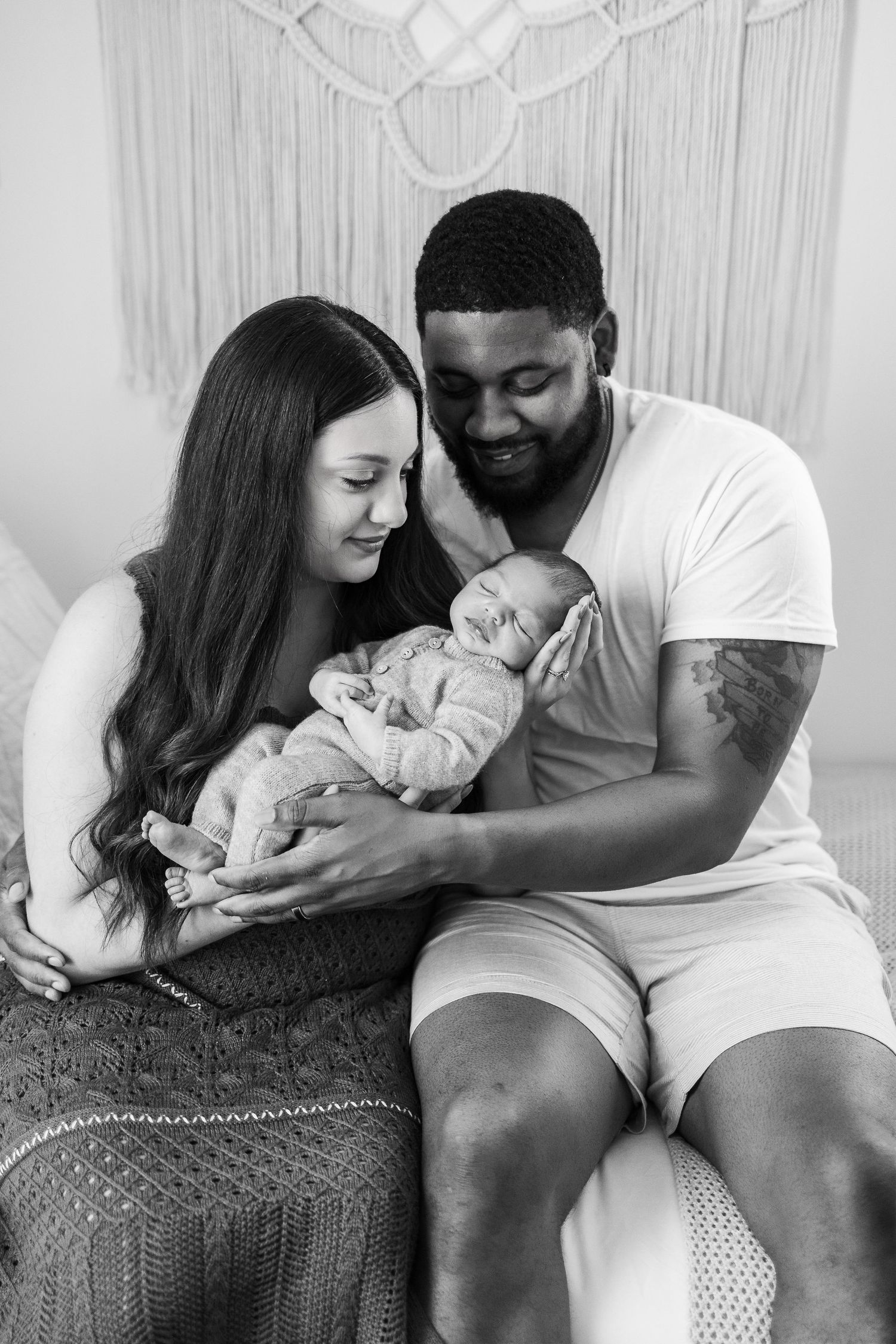 A loving couple gently holds their newborn baby in black and white portrait against macrame backdrop.
