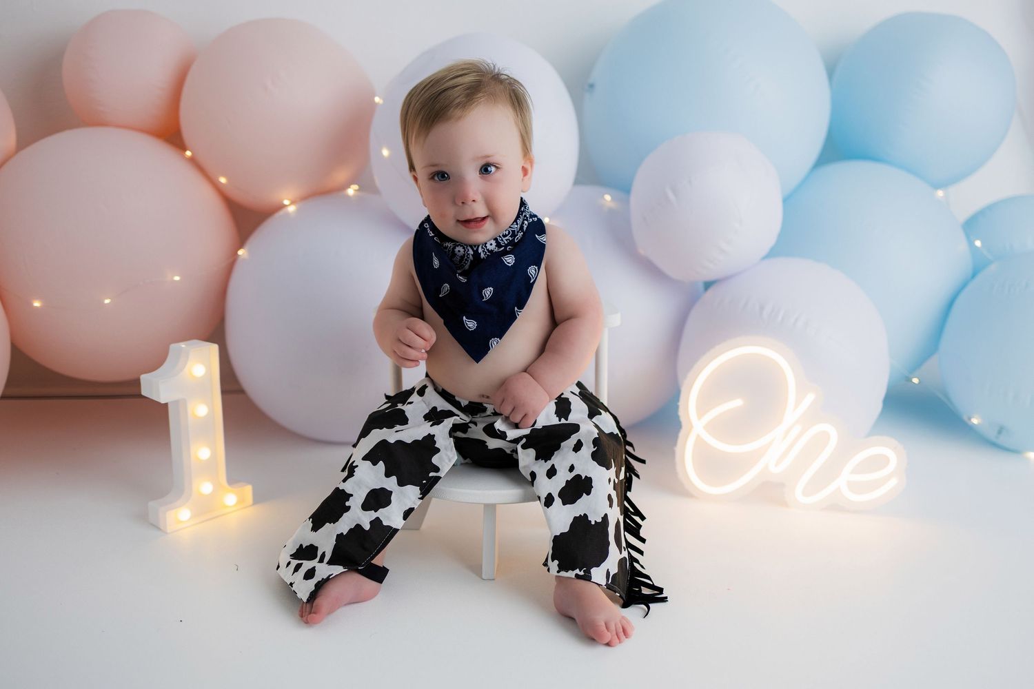 First birthday celebration setup with illuminated number one, pink and blue balloons, and baby sitting on floor.