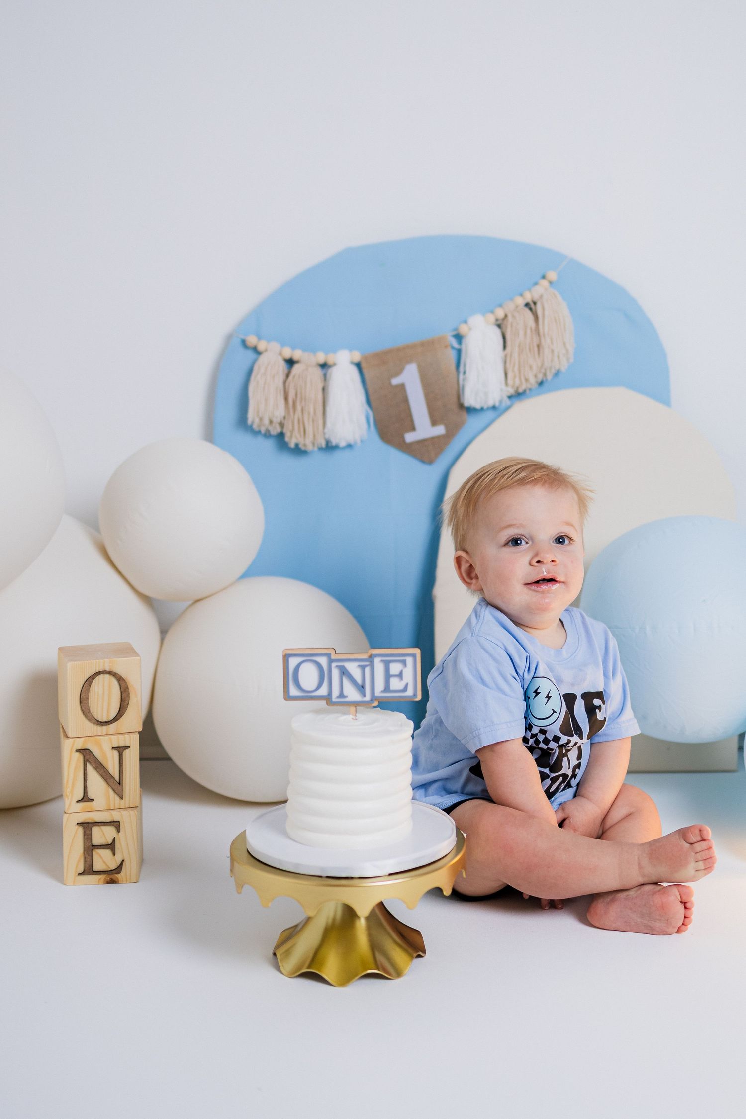 First birthday cake smash photo session setup with white and gold letters, white cake, and blue backdrop.