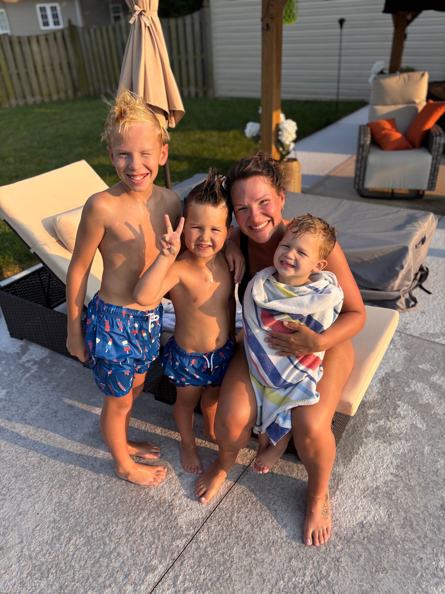 A woman smiles with three young children in swimwear by a backyard pool on a sunny evening.