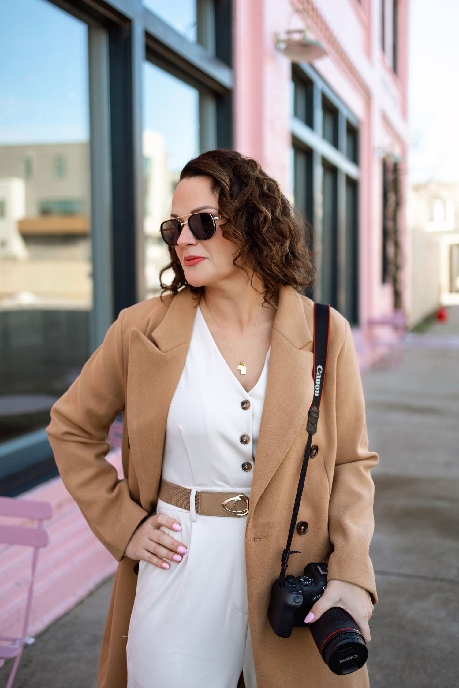Stylish woman in white dress and camel coat poses in four similar shots outside a building with pink accents.