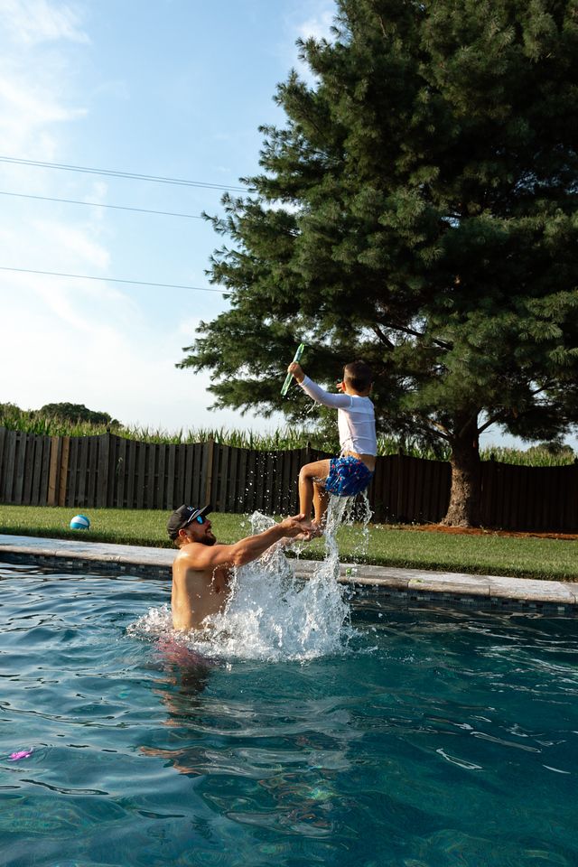 Adult tossing a child into the air above a backyard swimming pool on a sunny summer day.