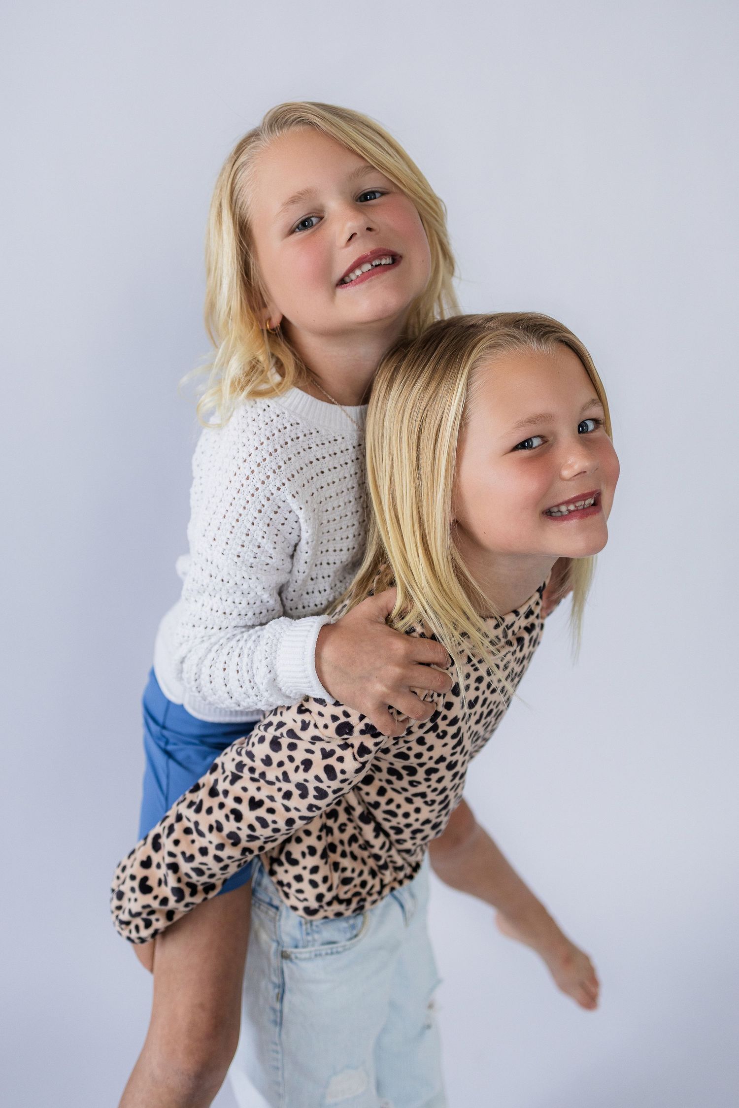 Two blonde children playing and laughing together, one giving a piggyback ride to the other against a gray background.