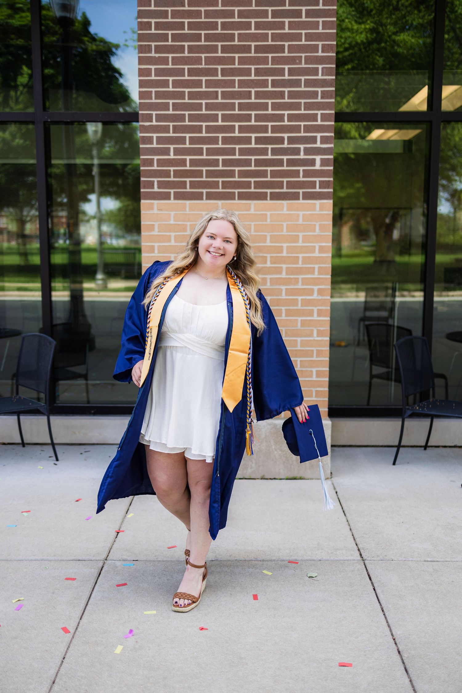 Graduate in white dress and blue gown smiles while posing outside building with glass windows.