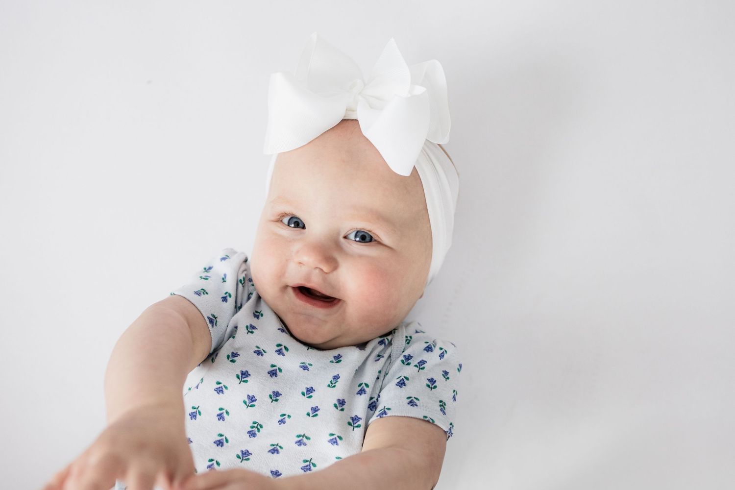 Baby wearing white bow headband and floral print outfit against white background.