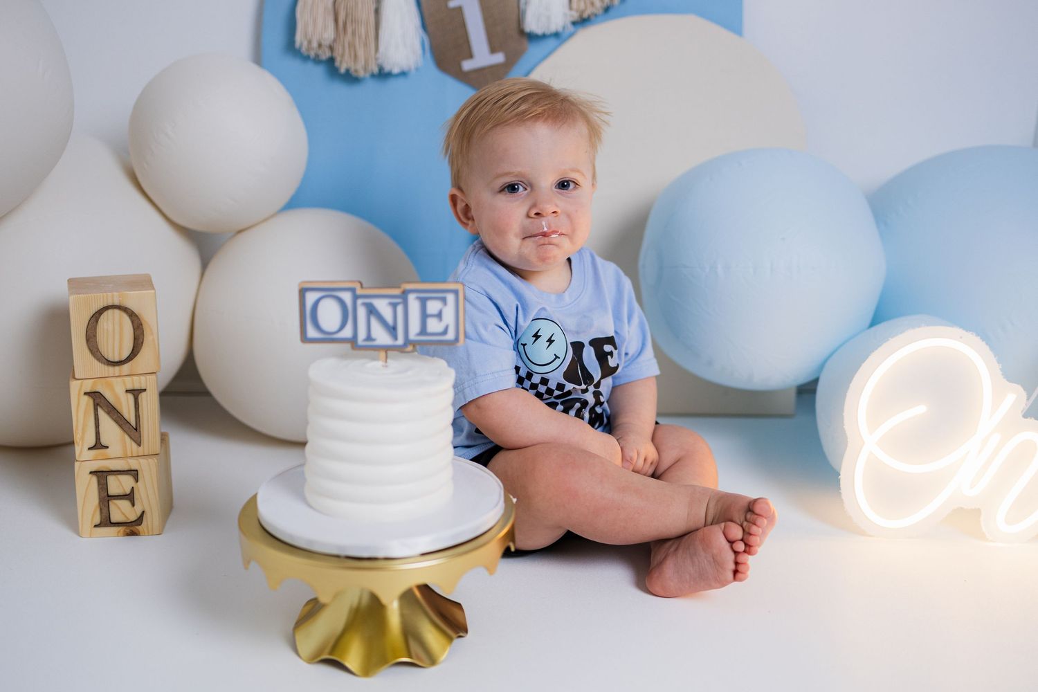 First birthday celebration setup with wooden blocks spelling ONE, white cake on gold stand, and blue and white balloon decorations.