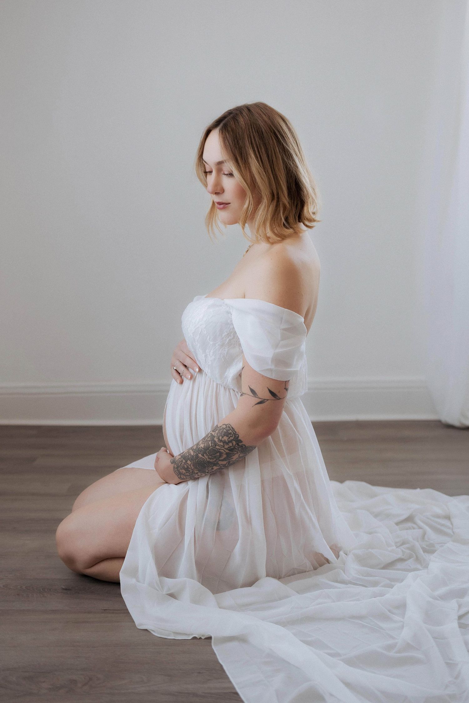 Artistic maternity portrait with flowing white dress on wooden floor.