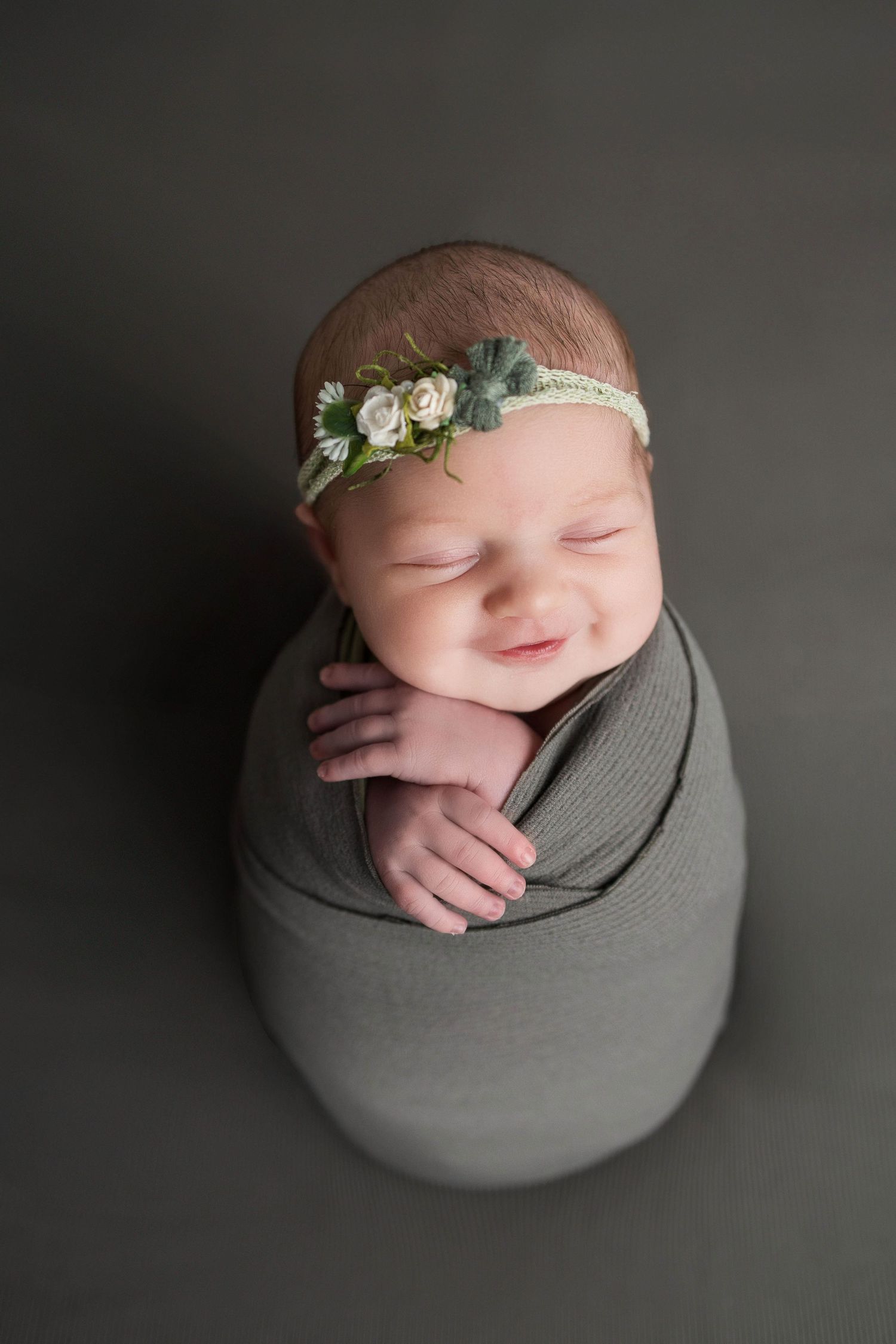 Newborn baby photographed curled up on dark grey background wearing delicate floral crown in artistic pose.
