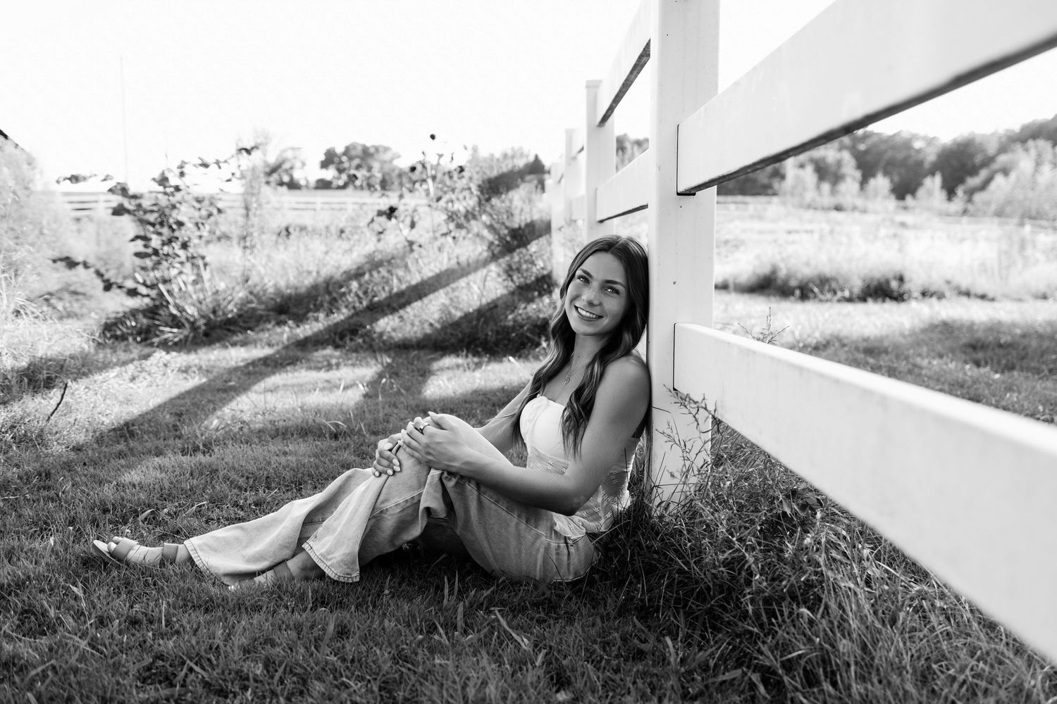 Black and white photo of a smiling young woman sitting on grass leaning against a white fence outdoors.