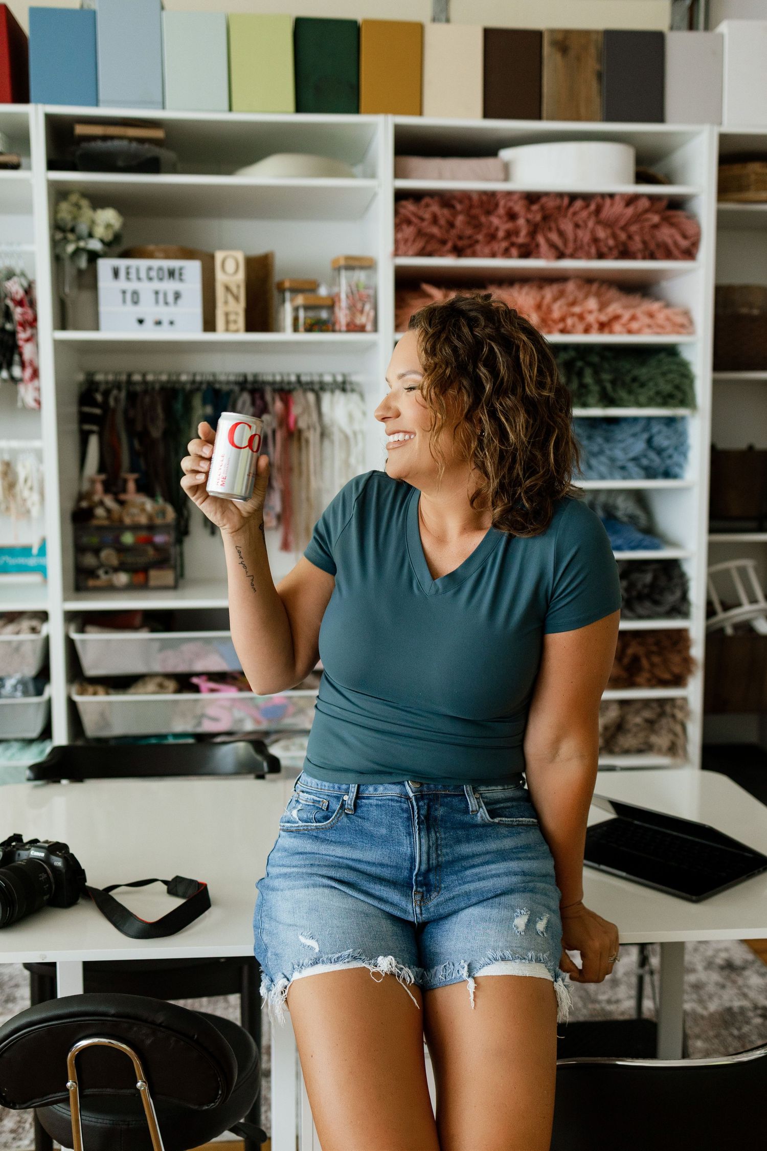 Woman smiling in craft studio holding mug, sitting on desk with fabric shelves and Welcome sign in background.