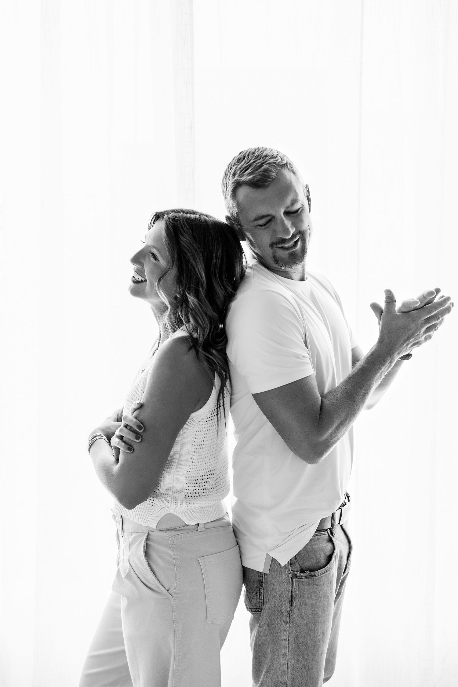 Black and white photo of a smiling couple standing back to back in casual white outfits against a bright curtain backdrop.
