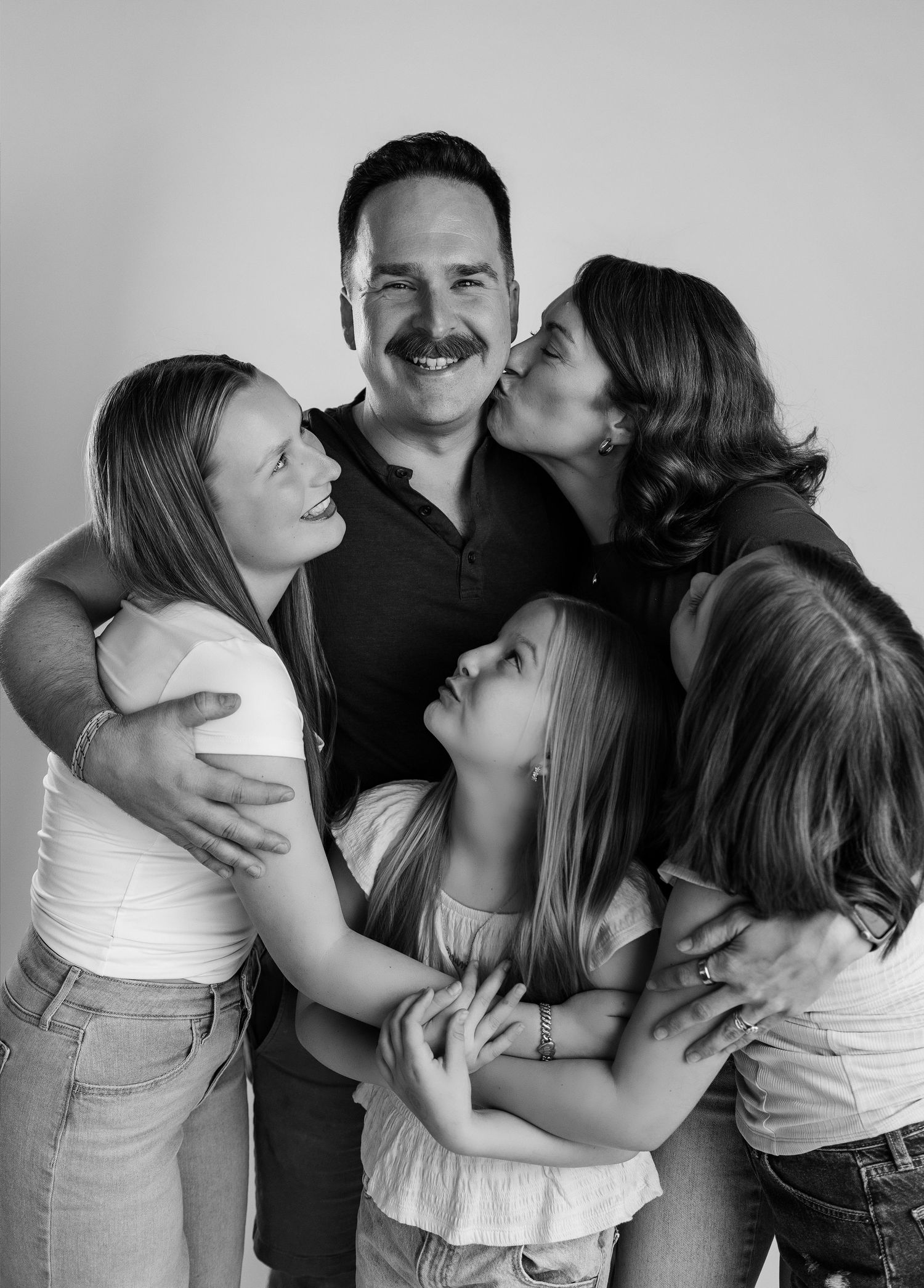 Black-and-white portrait of a father being hugged and kissed by his family during a personality session