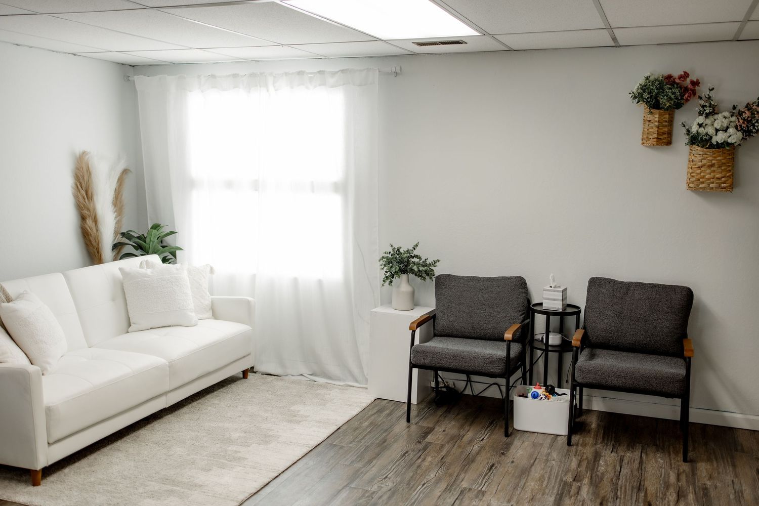Modern minimalist living room with white sofa, grey chairs, wood flooring and natural light from large windows.