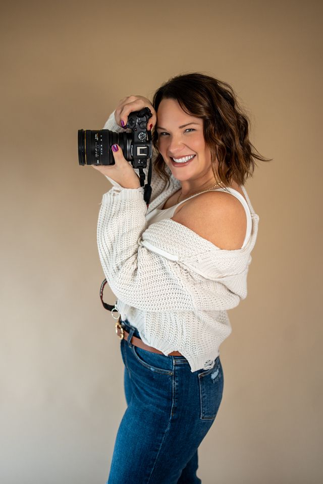 Photographer in white sweater and jeans smiling while taking photos with professional camera.