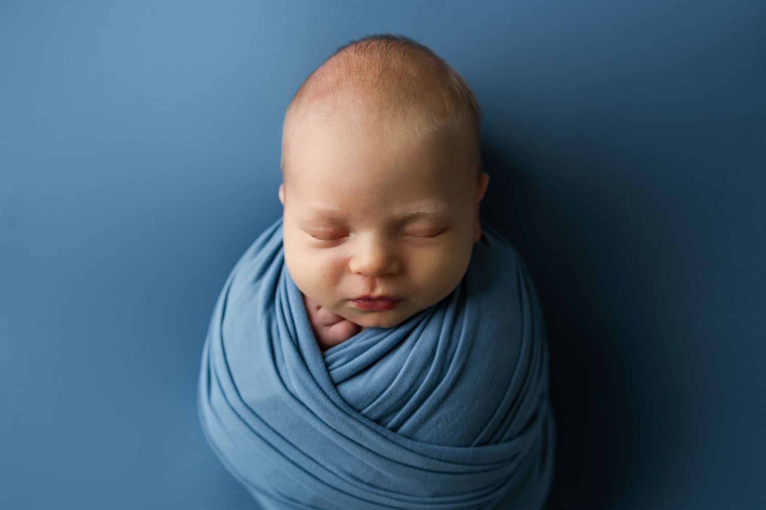 A sleeping newborn baby swaddled in soft blue fabric against a navy blue background.
