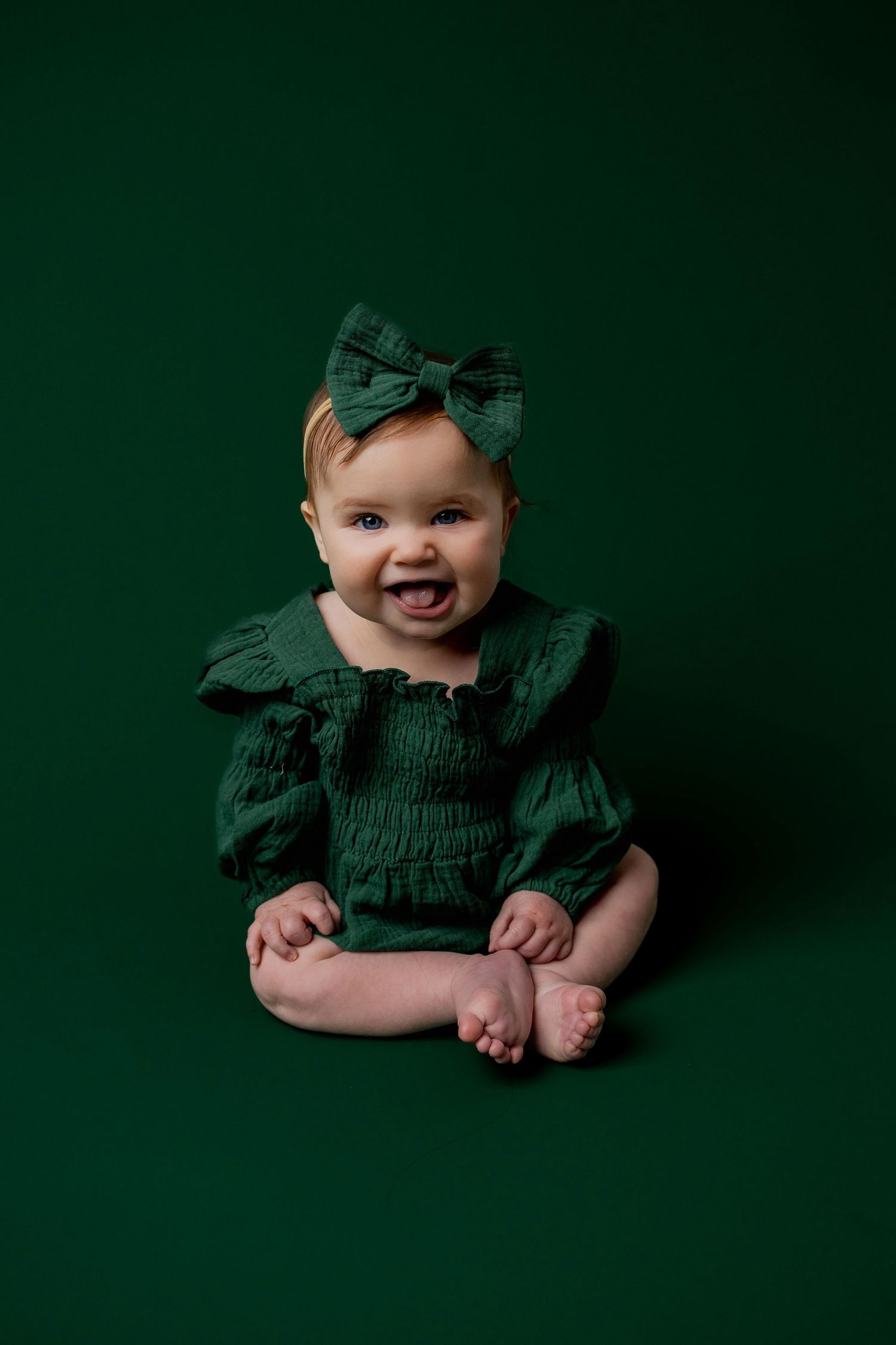 Professional baby portrait against dark green backdrop wearing matching green ruffled outfit and bow.