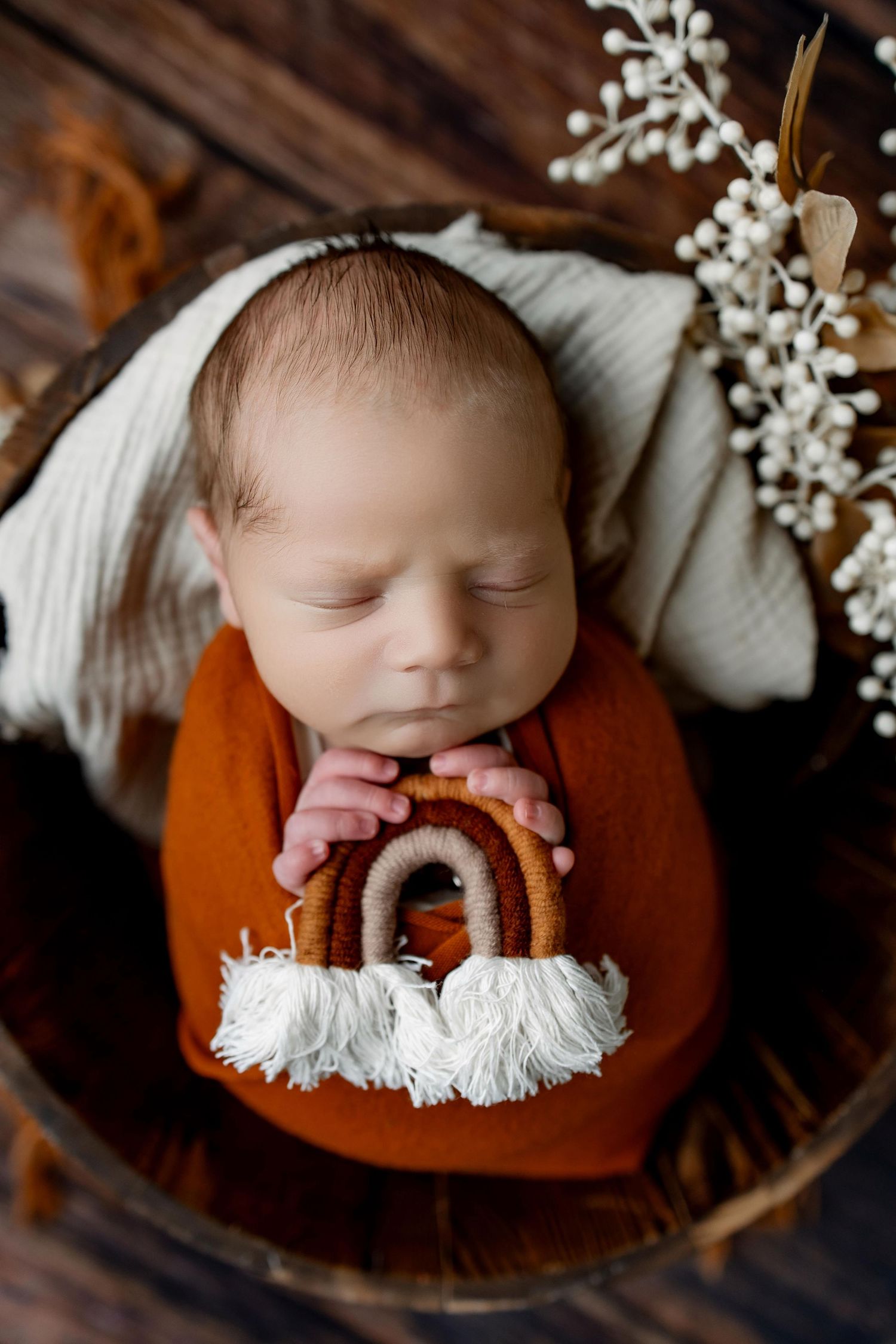 Sleeping baby in rust orange outfit holding wooden rainbow toy with white tassels on dark wooden surface.