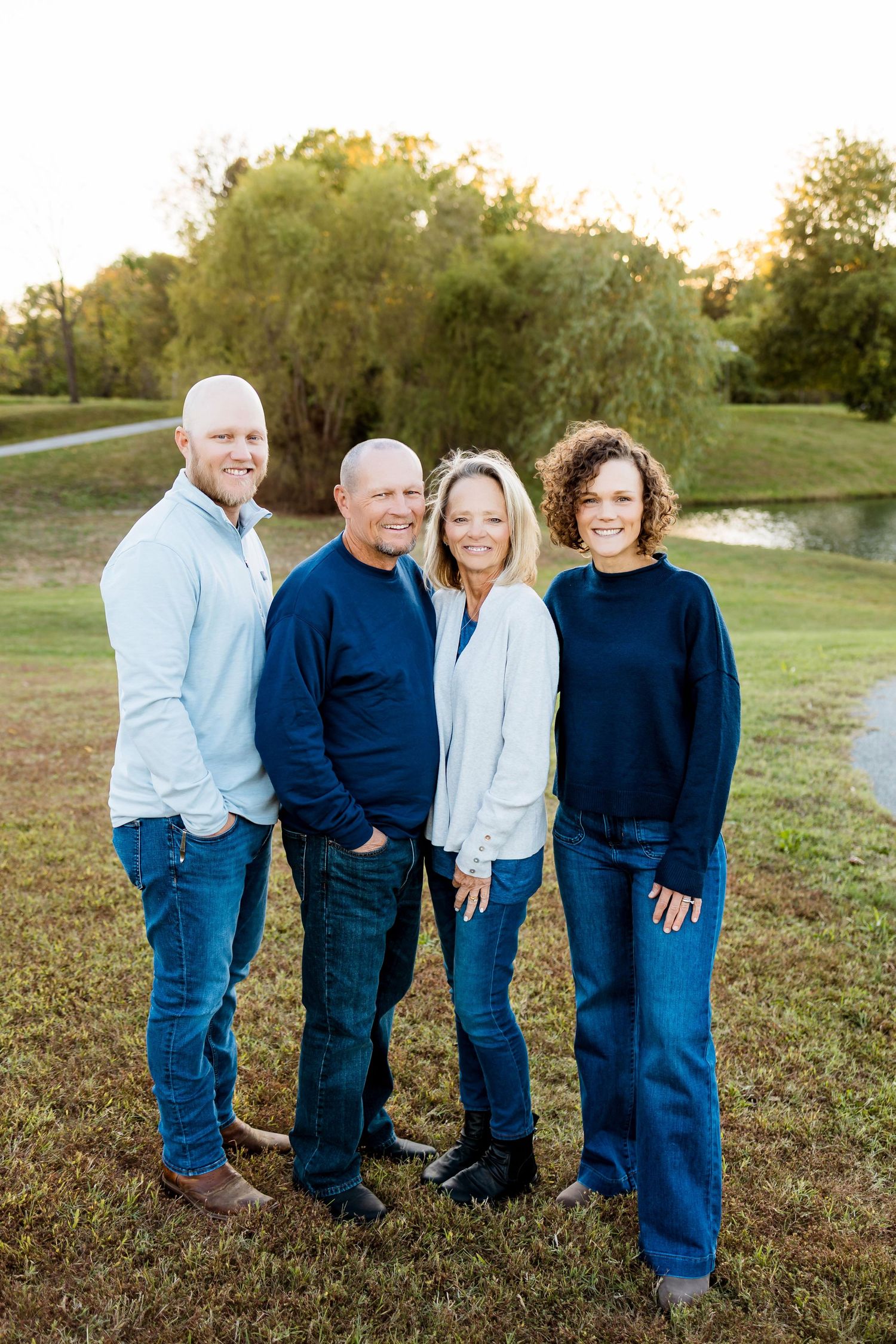 A family wearing coordinating blue and white outfits poses together outdoors in a grassy field at sunset.