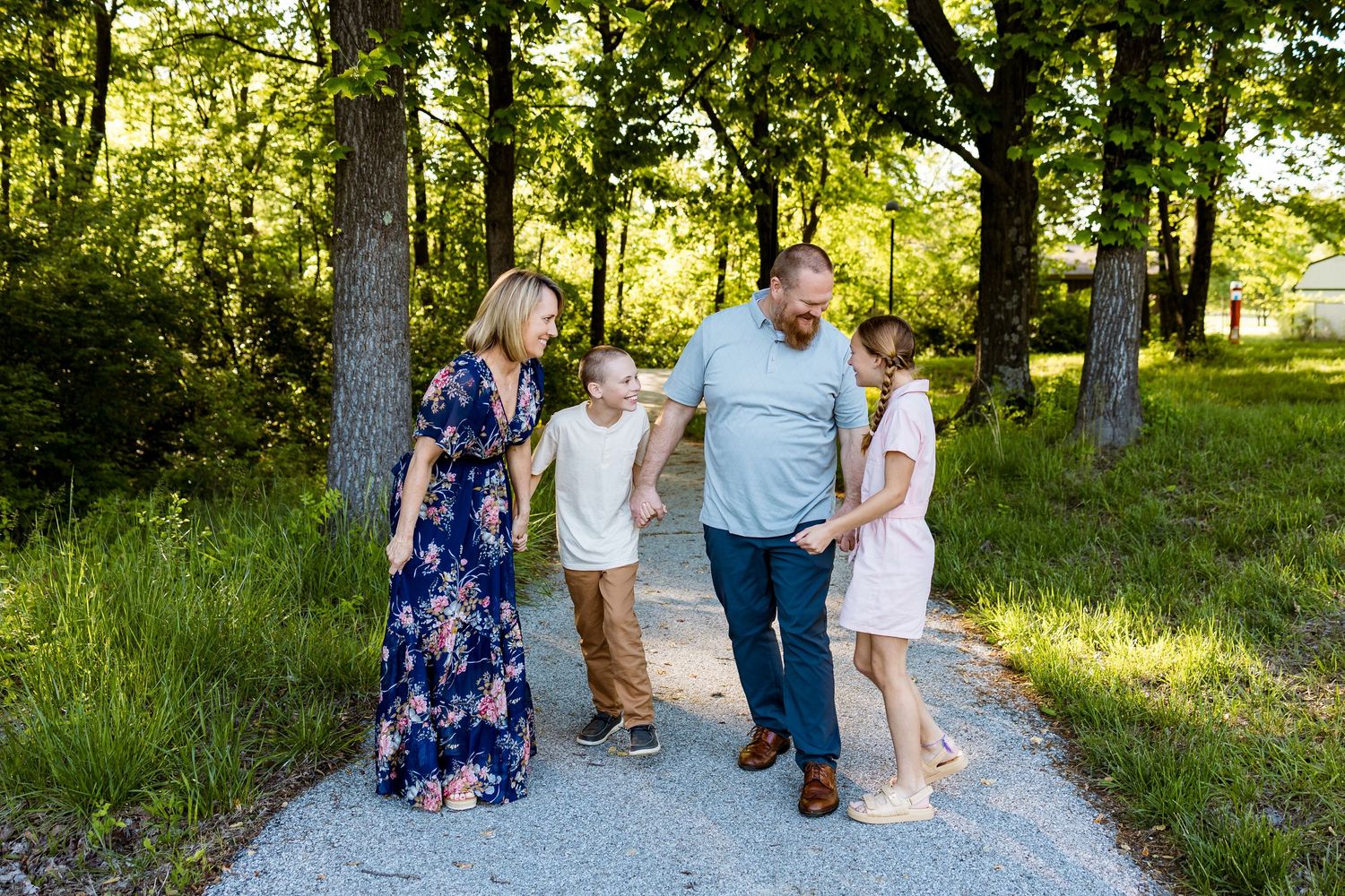 A family walks together along a winding gravel path through a sunlit wooded area.