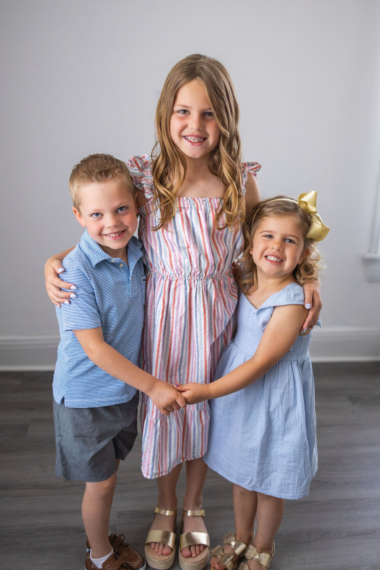 Three children in coordinating blue and pink outfits pose together against a gray wall on hardwood floors.