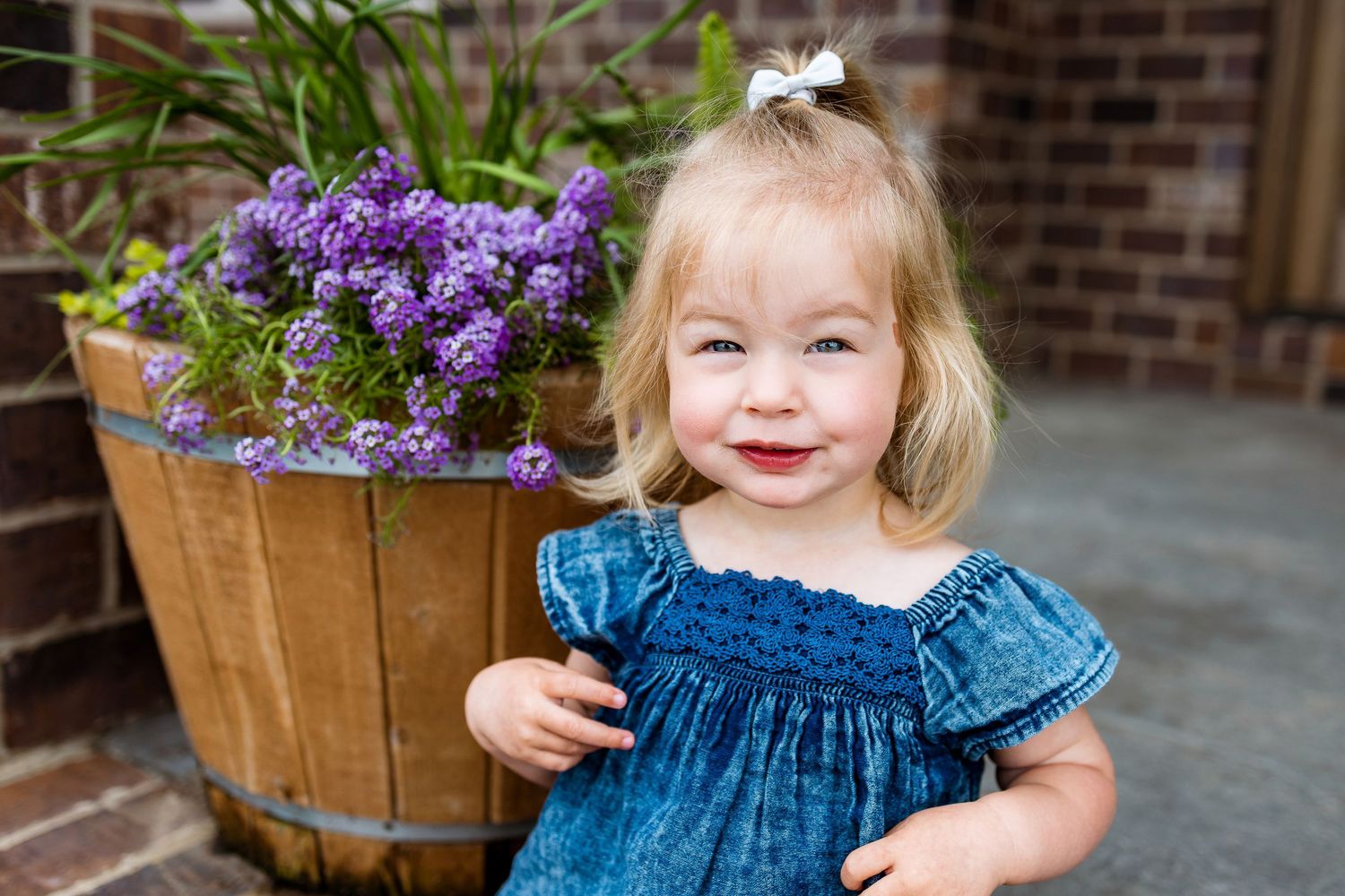 A smiling child in a blue dress stands next to a wooden barrel planter with purple flowers.