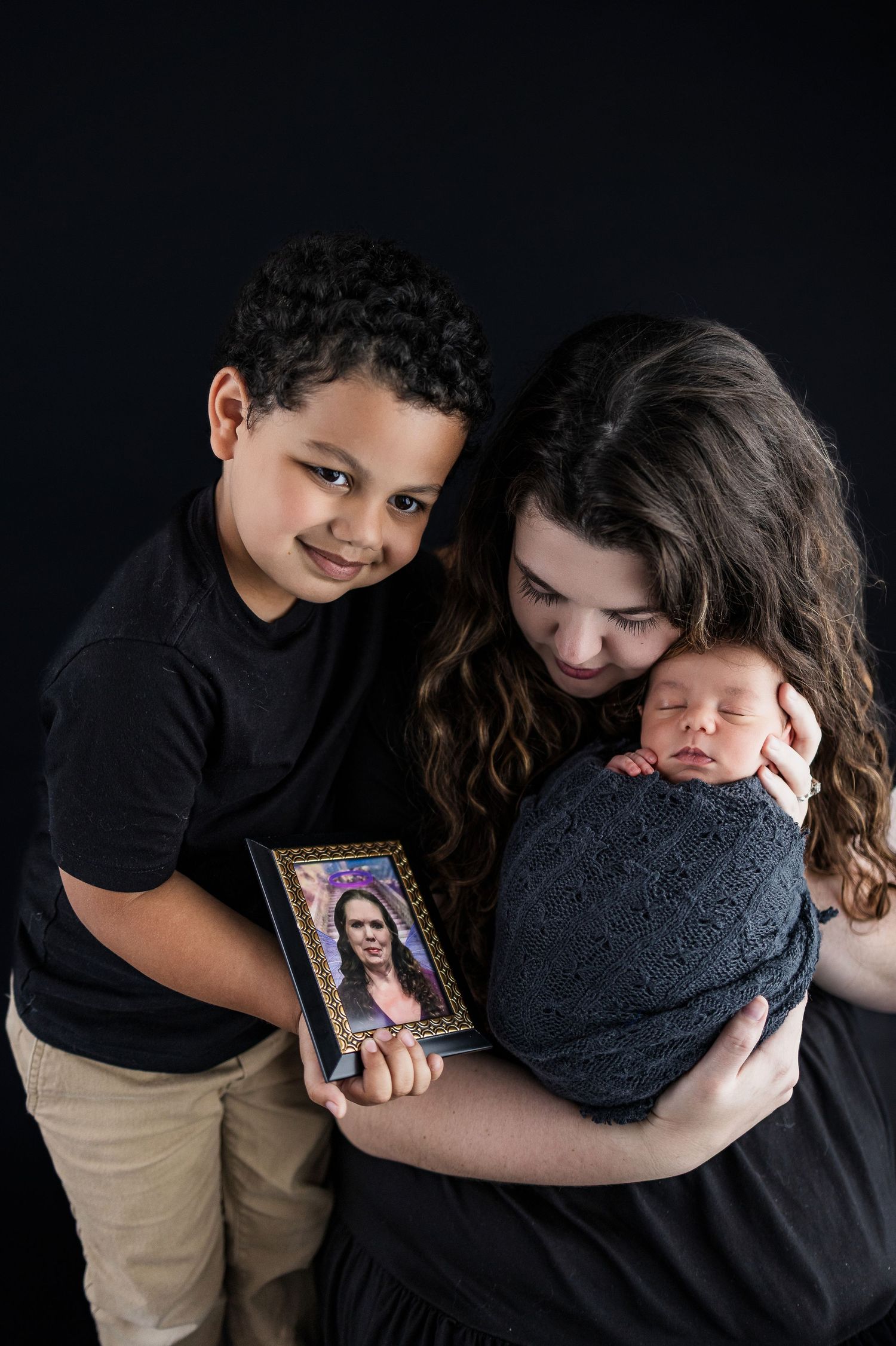 A family poses together against a black backdrop while holding a framed photograph and newborn baby.