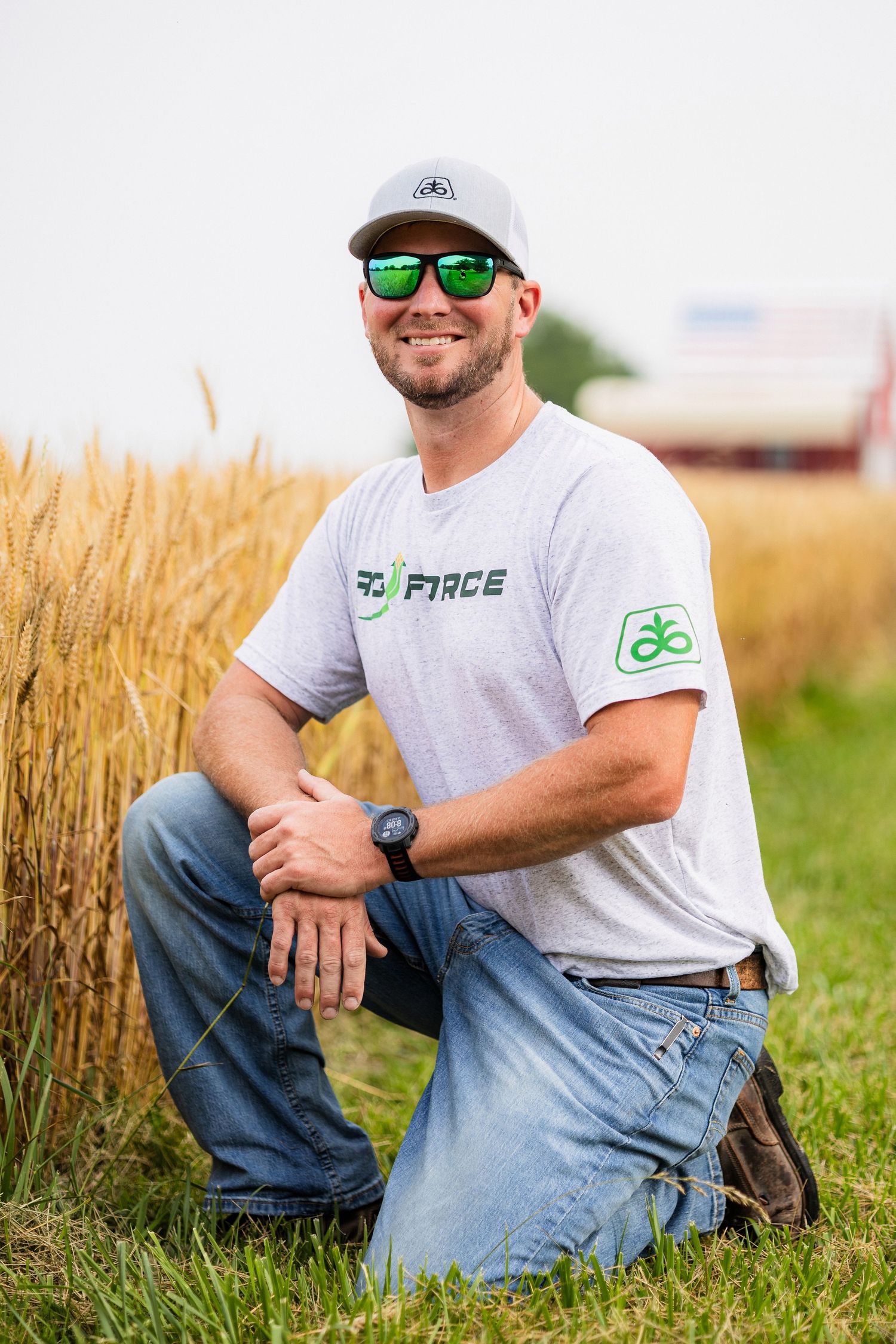 Person in casual attire sitting in agricultural field with sunglasses and baseball cap.