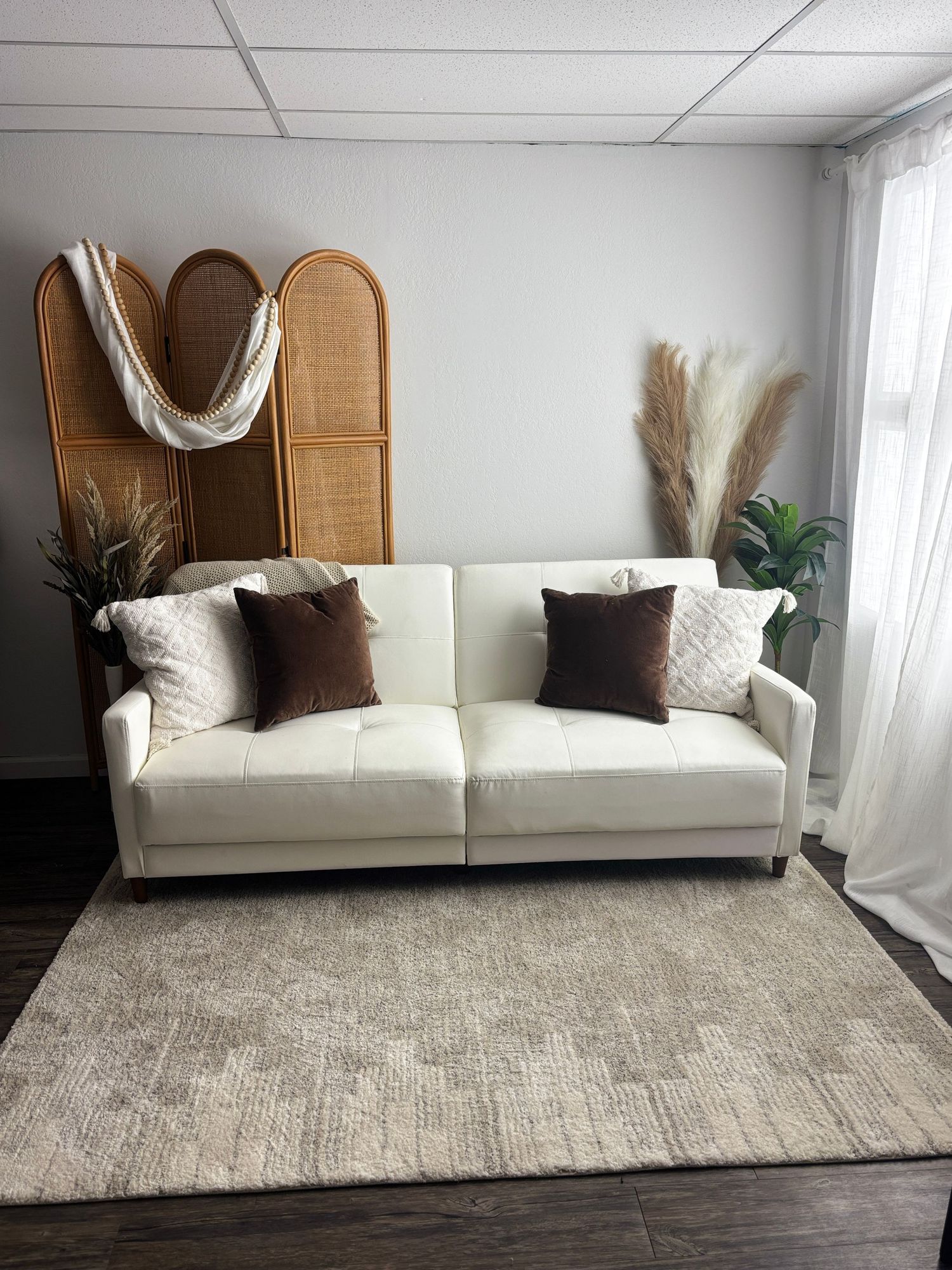 Modern white sofa with brown throw pillows against rattan room divider, styled with pampas grass and area rug in neutral tones.