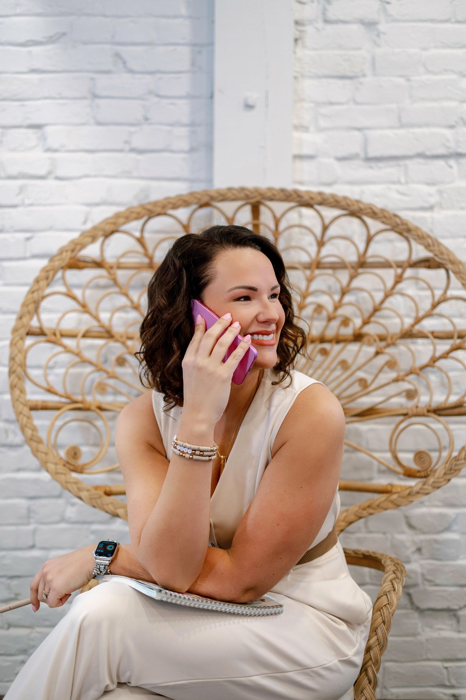 A person in a white dress laughs joyfully while sitting in an ornate rattan peacock chair against a white brick wall.