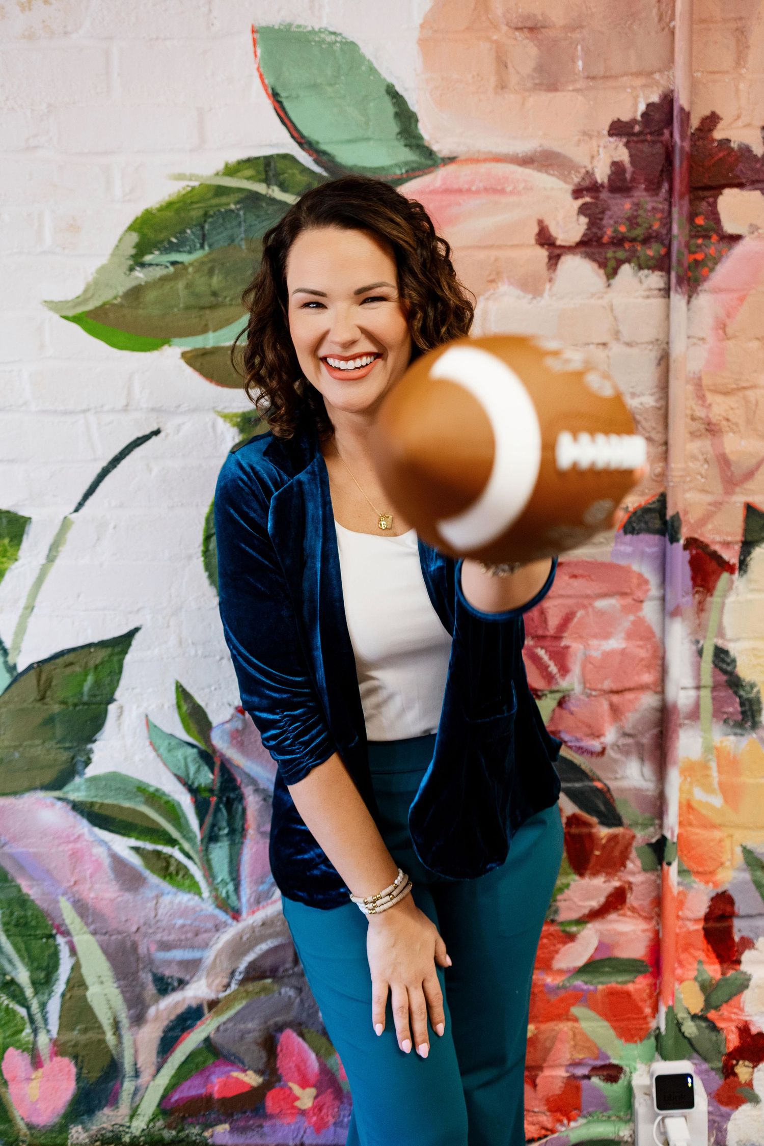 A person in a navy blazer and white top poses playfully against a colorful painted wall with green plants.