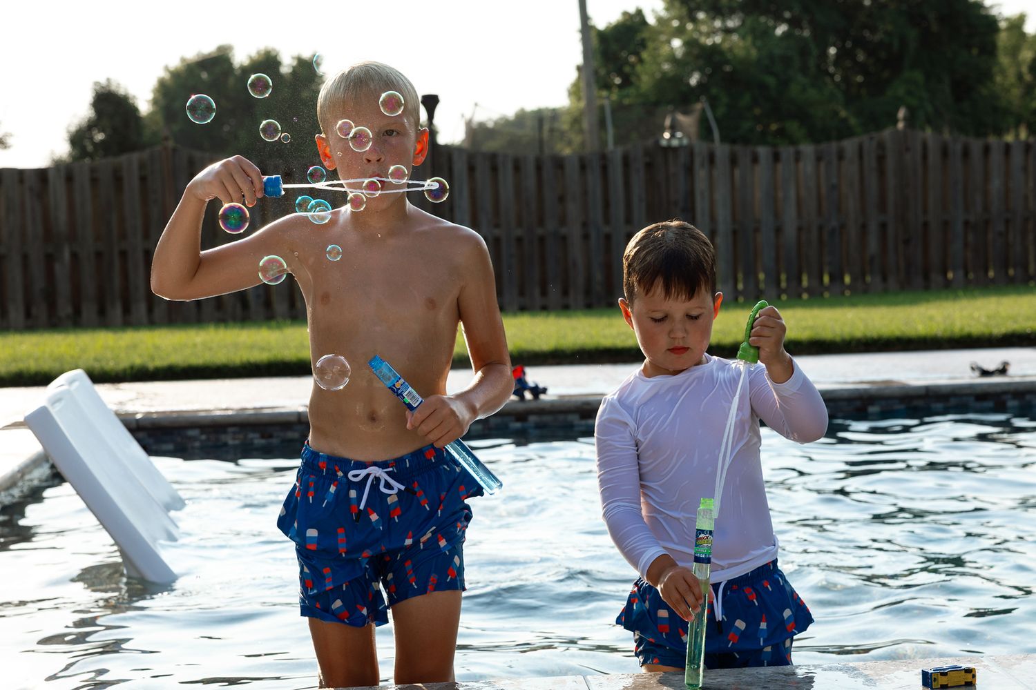 Two boys playing with bubble wands in a backyard pool on a sunny day.