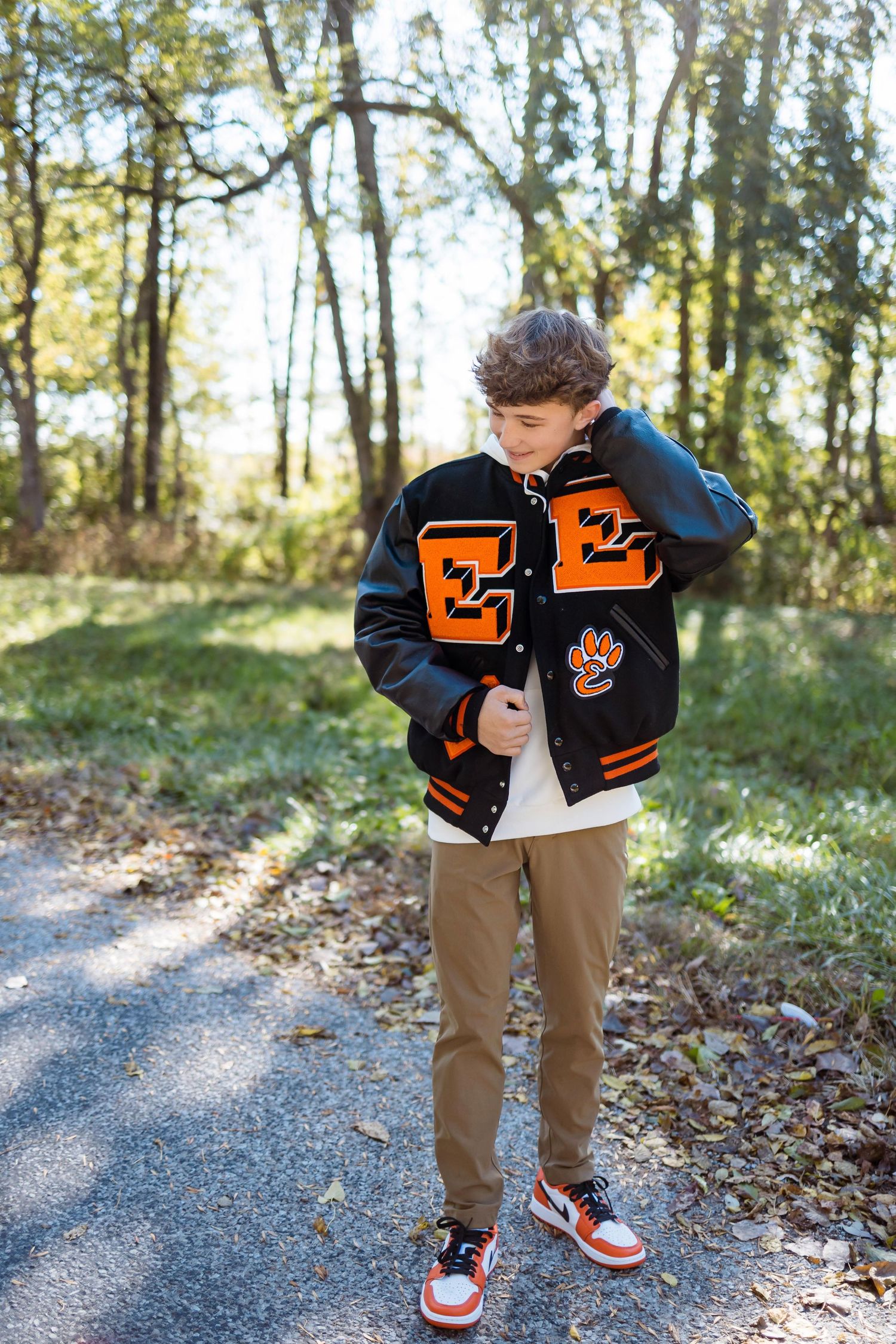 A person wearing a black varsity jacket with orange letters and khaki pants stands on a leaf-covered path in autumn.