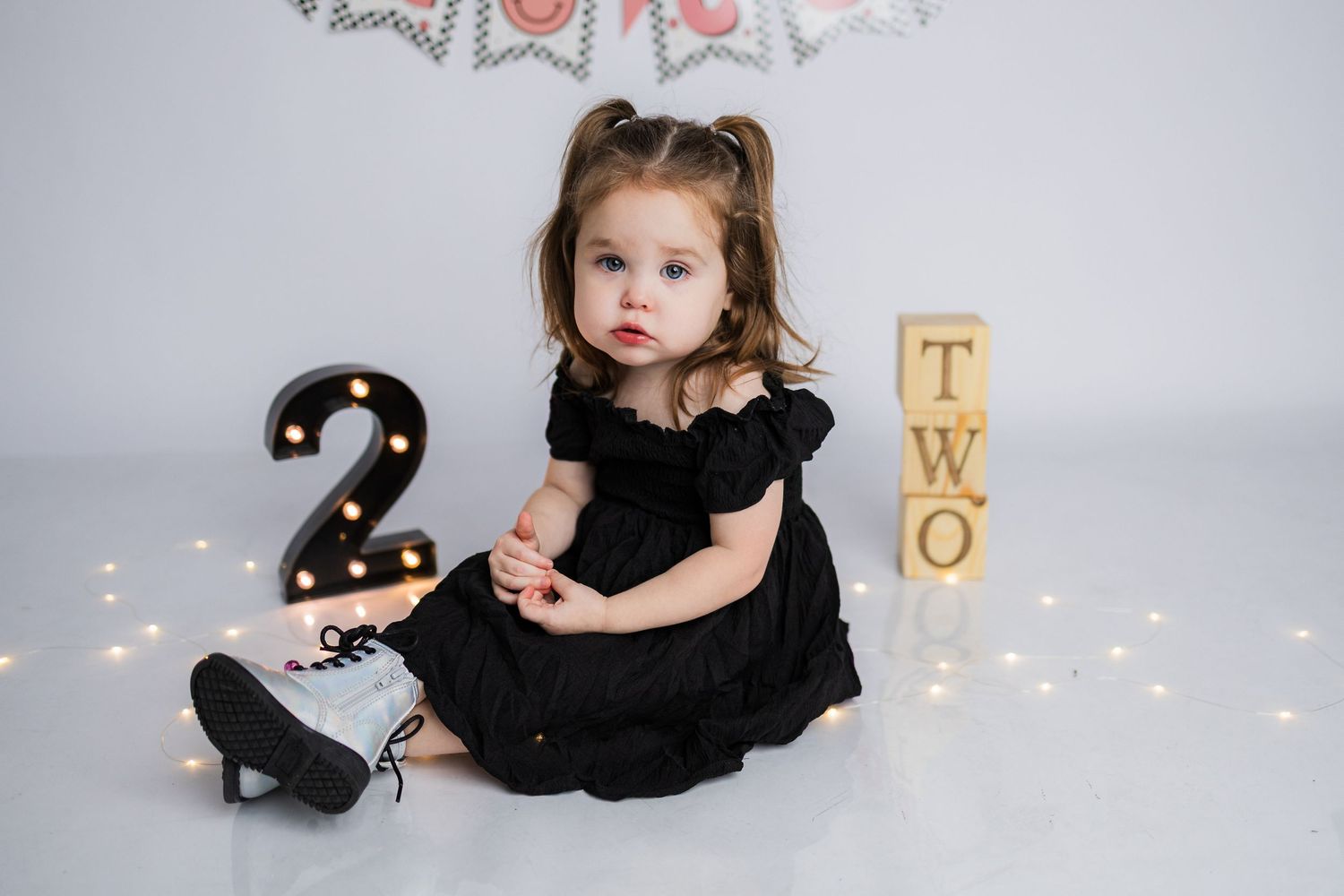 Small figure in black dress poses on light backdrop with glowing string lights and festive decor.