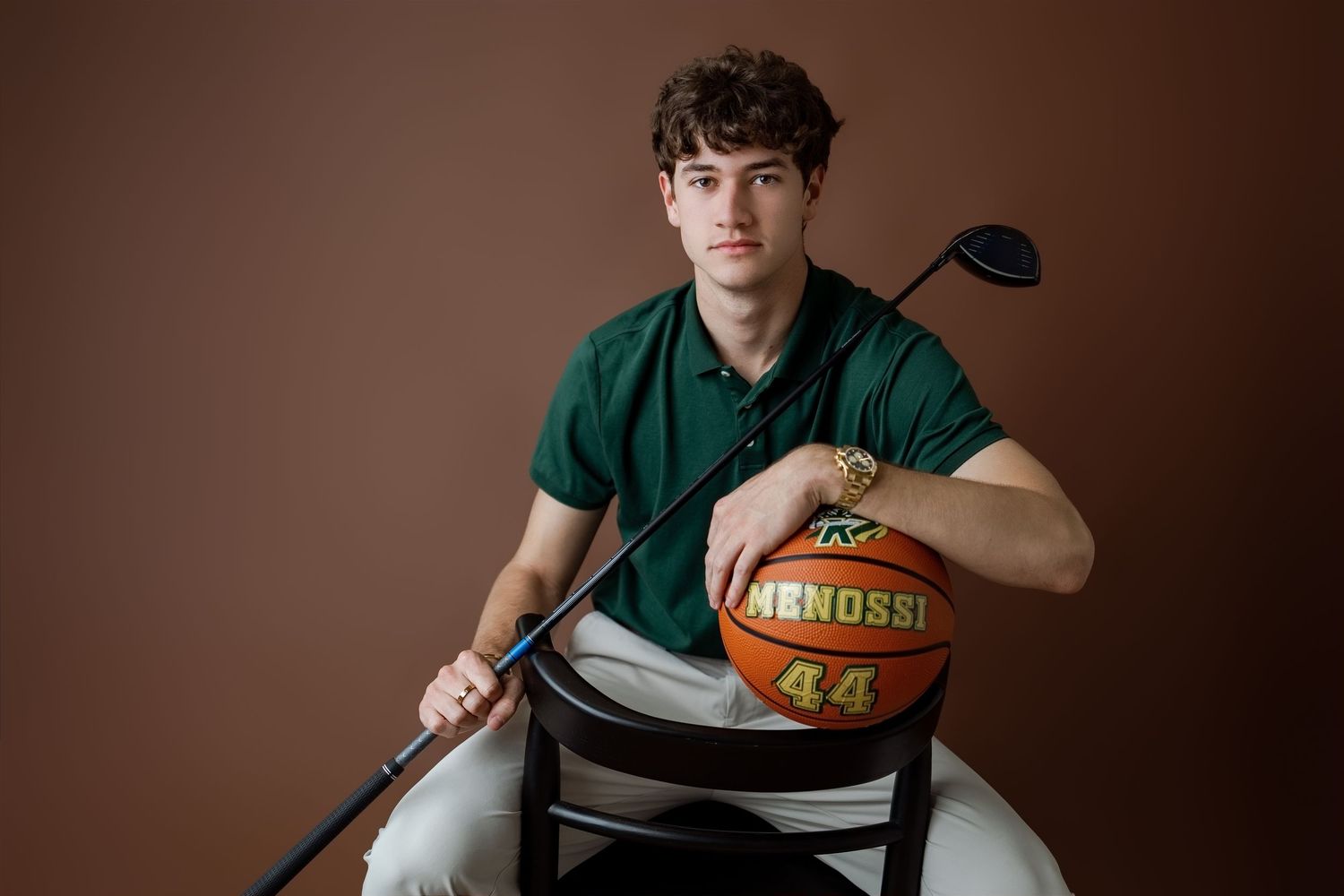 Teen boy in green polo holds golf club and basketball with number 44, posed on chair against brown background.